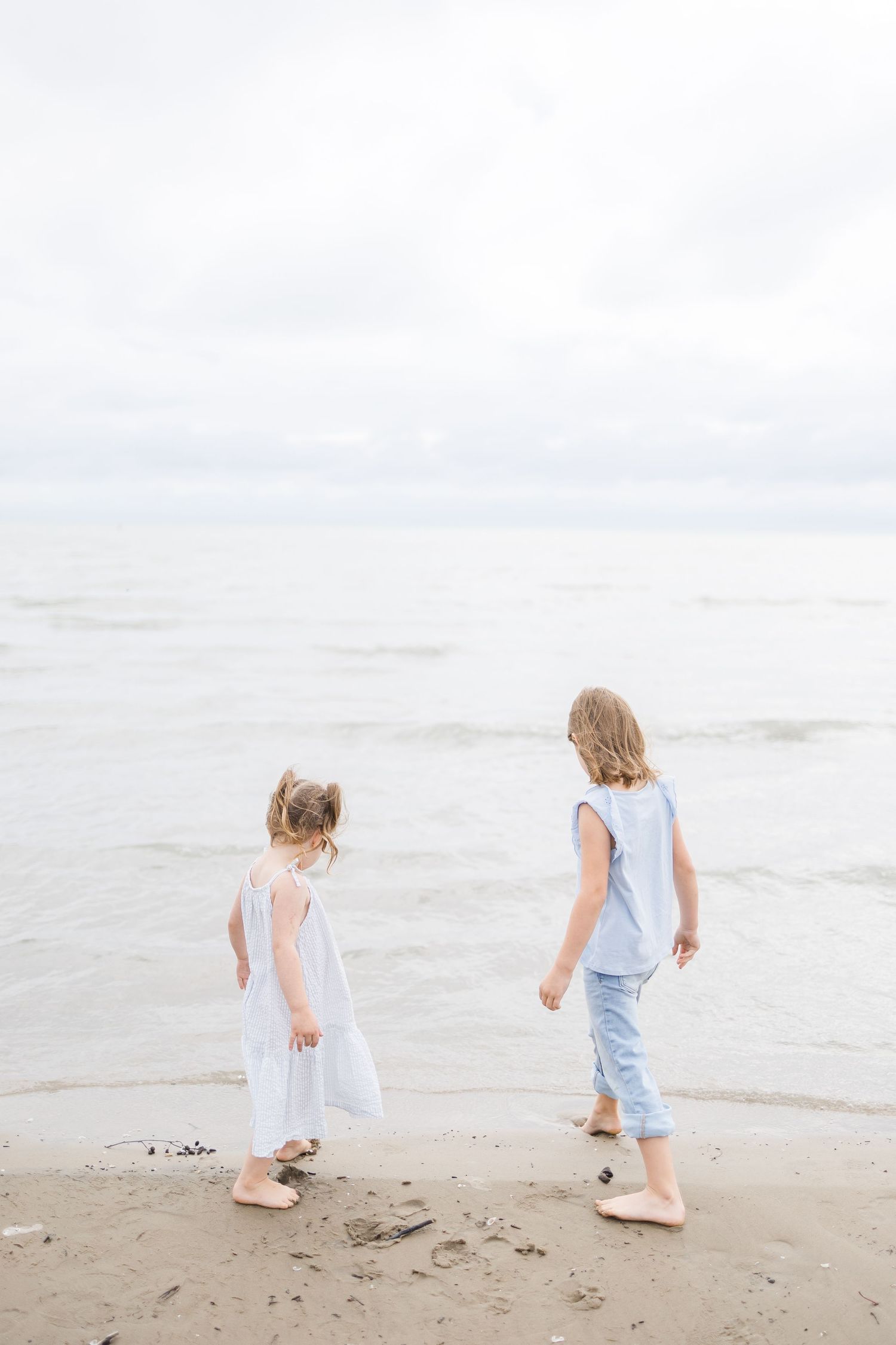 Two young children in light colored clothing walking together on a sandy beach by the ocean.