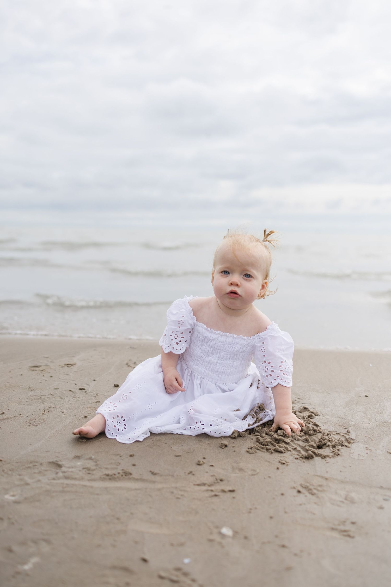 Baby in white lace dress playing in the sand at the beach.