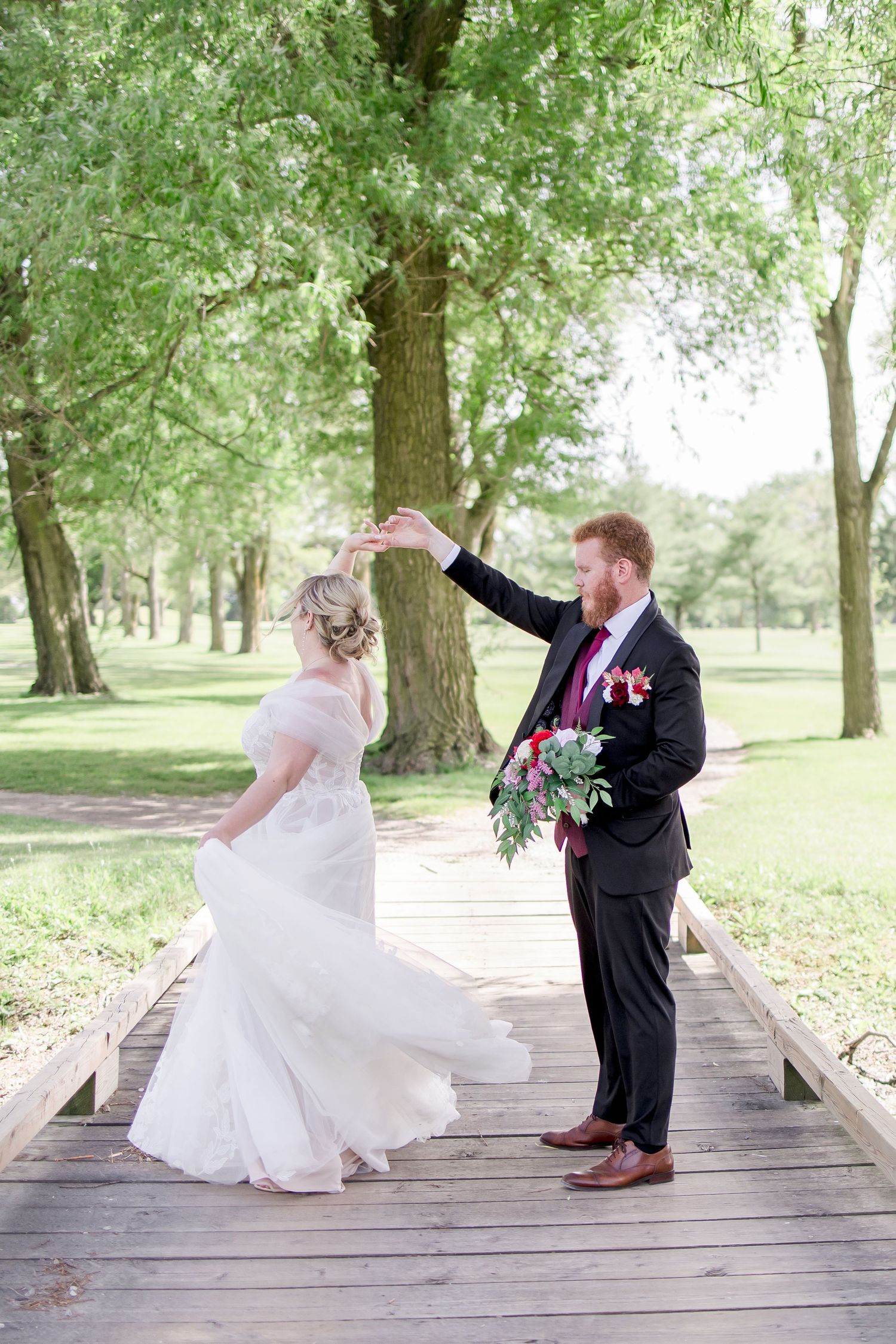 Playful wedding couple poses on wooden bridge in park setting.