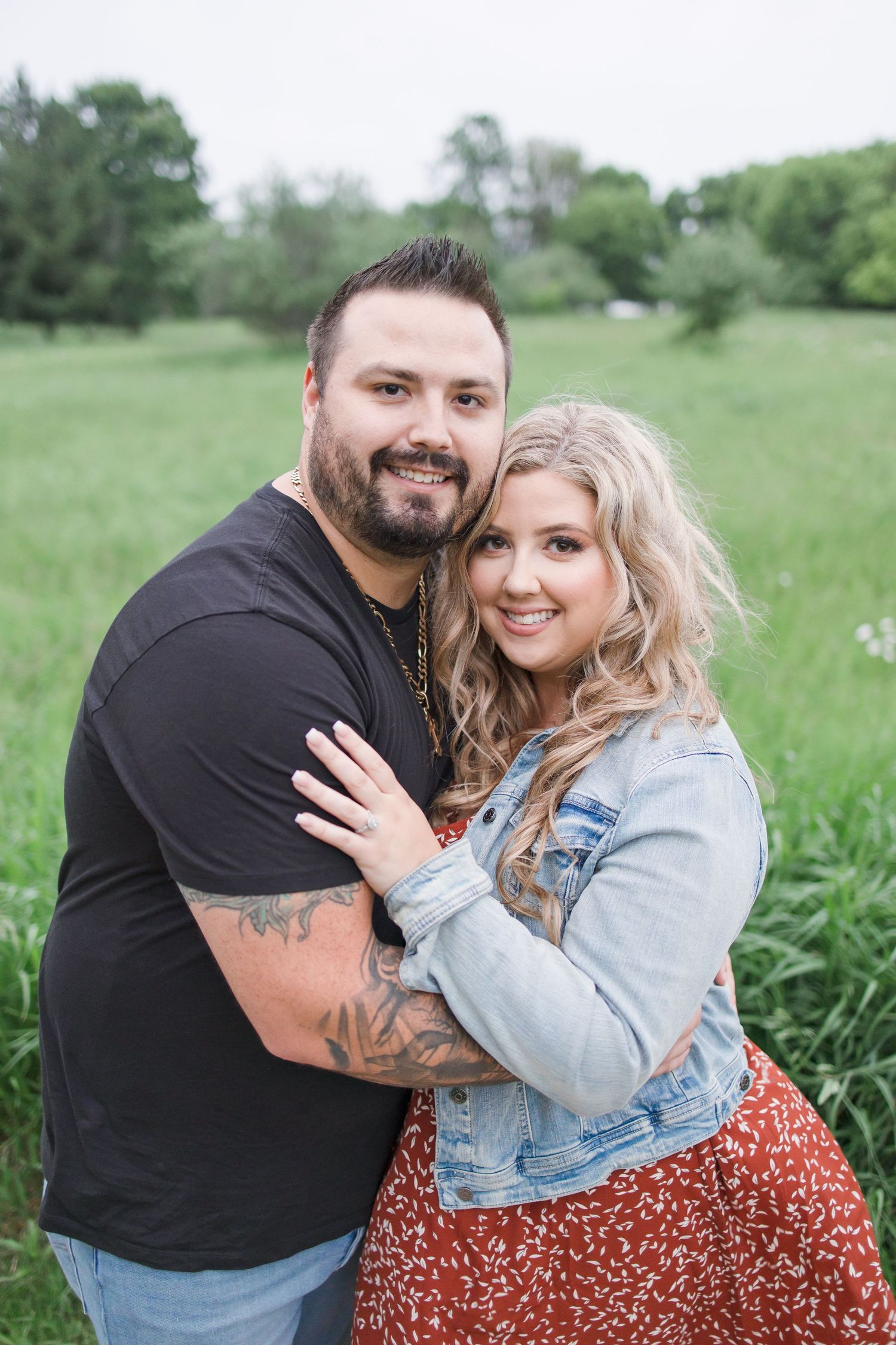 A couple poses together outdoors in a green field wearing casual denim and black attire.