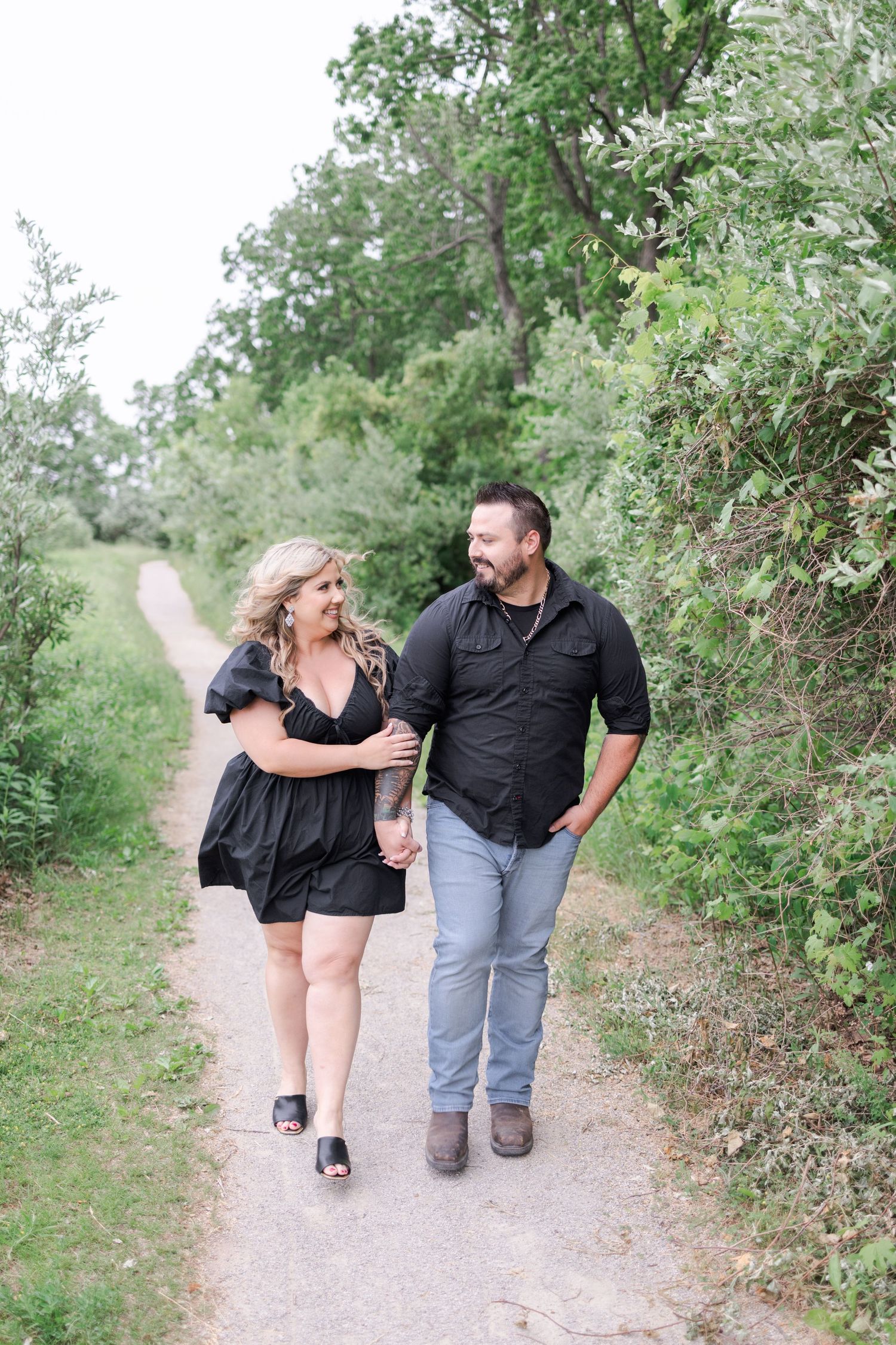 A couple walks together on a nature trail surrounded by green foliage on a sunny day.