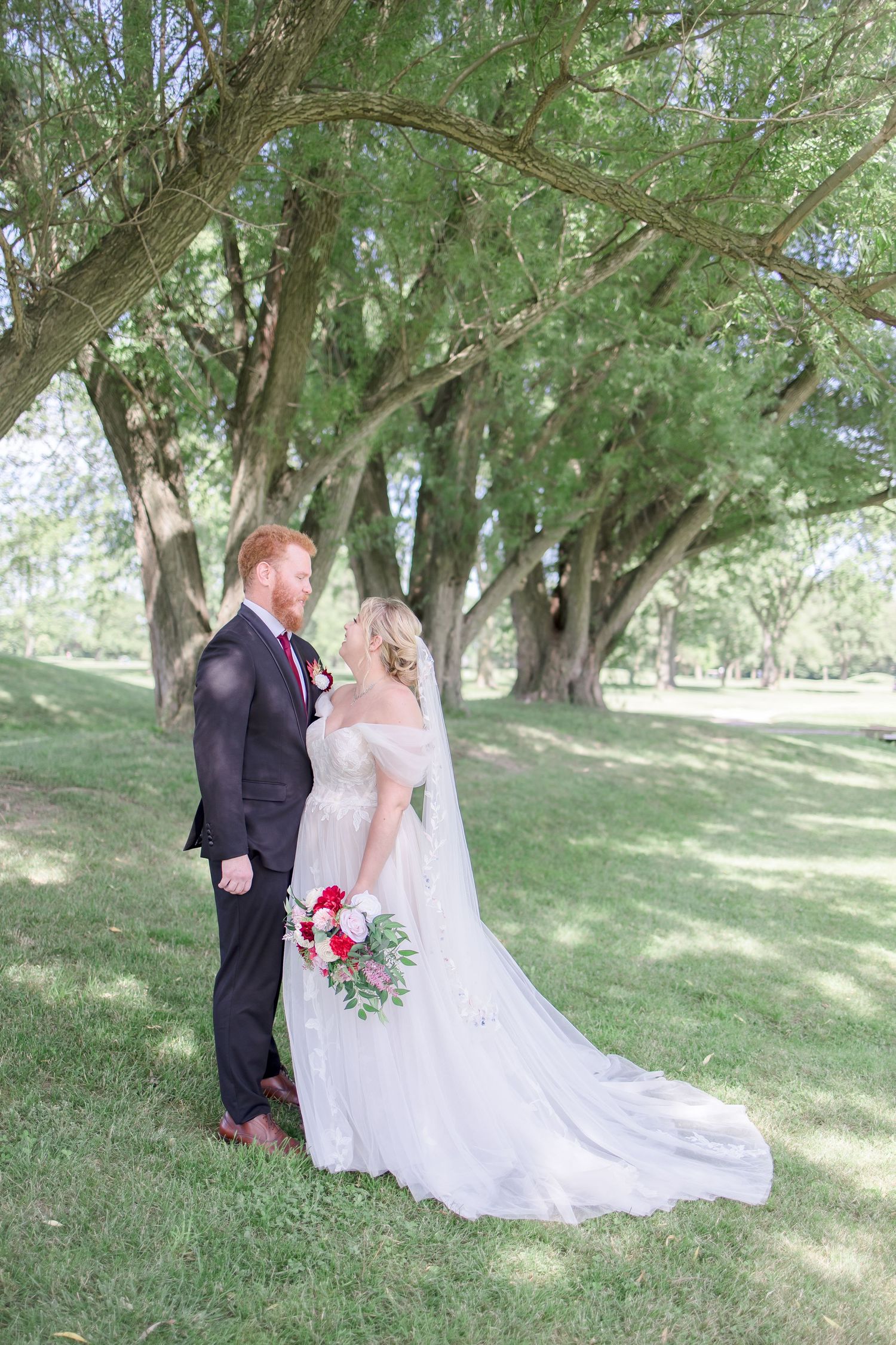 Elegant wedding couple poses under willow trees in outdoor garden setting.