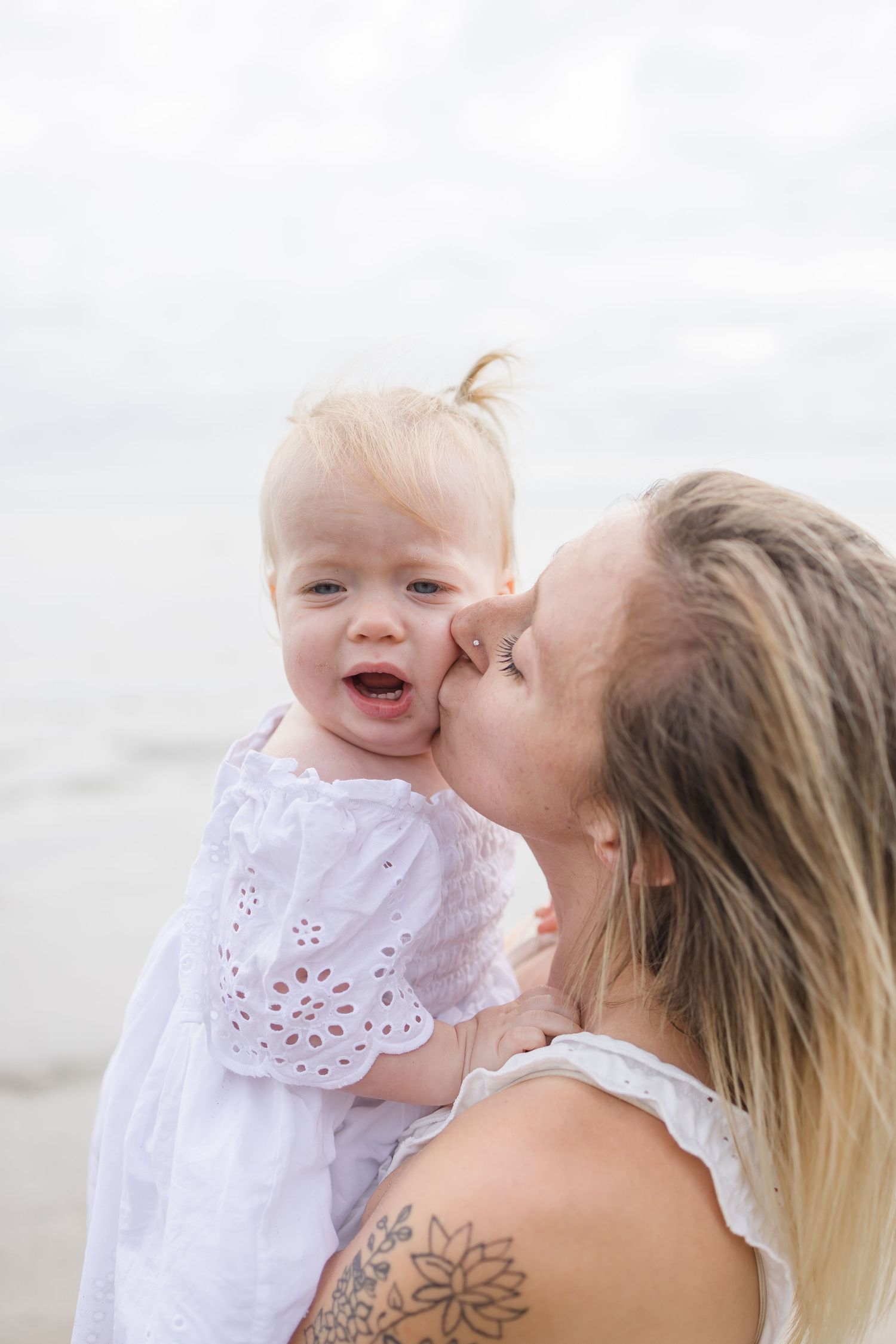 Mother and baby in white outfits sharing an intimate moment on the beach.