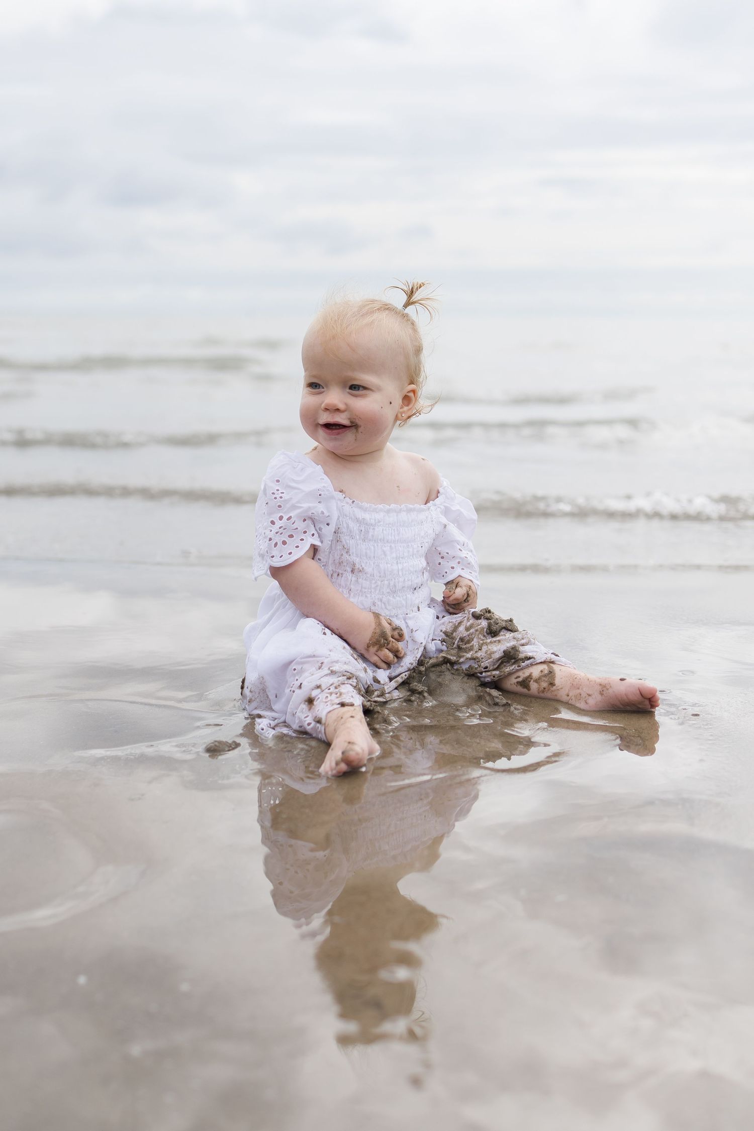 A toddler in white lace dress plays happily in shallow beach water.