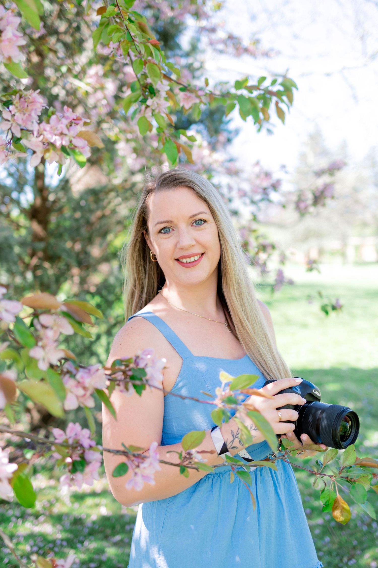 A photographer in a blue sundress stands among blooming spring cherry blossoms holding a camera.