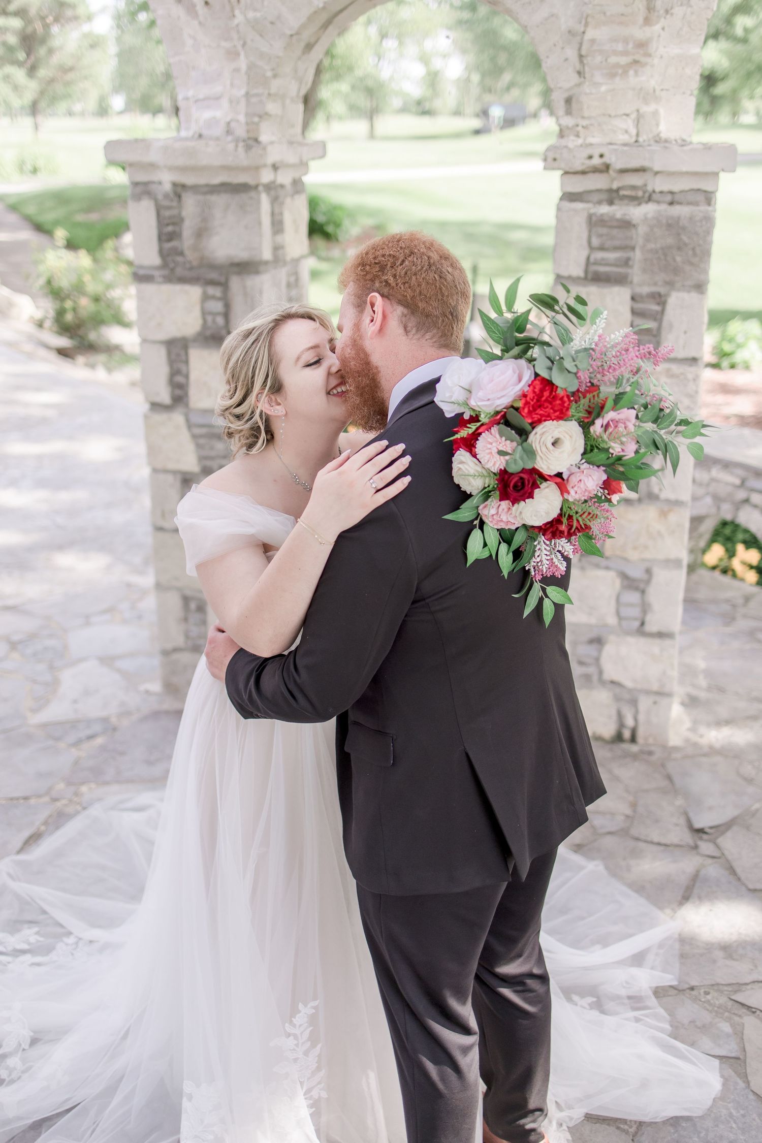 A romantic first look moment between newlyweds under a stone archway with red and white floral bouquet.