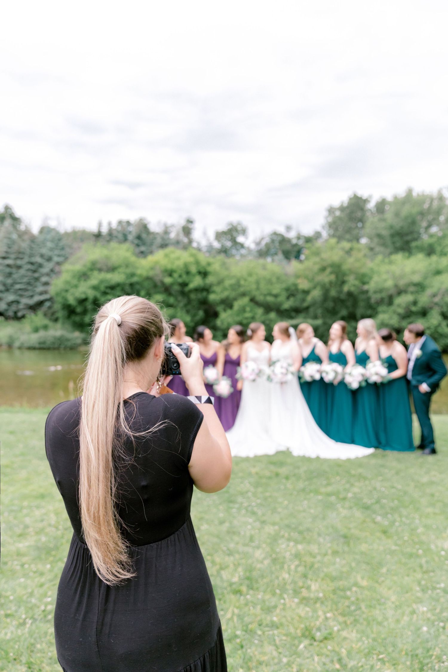 A photographer takes pictures of a wedding party by a lake with bridesmaids in teal dresses.