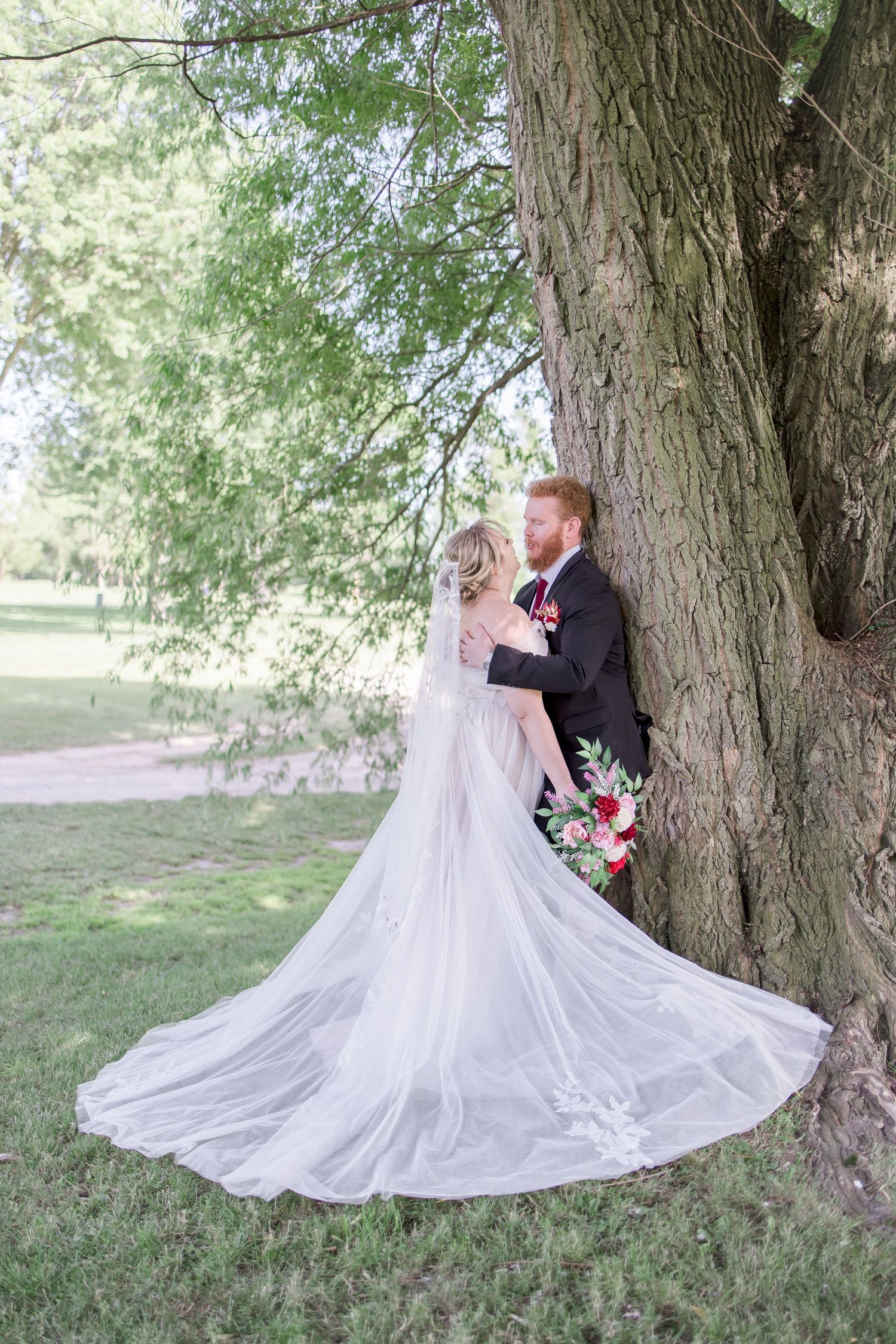 Flowing wedding dress train spreads beautifully against tree trunk in outdoor setting.