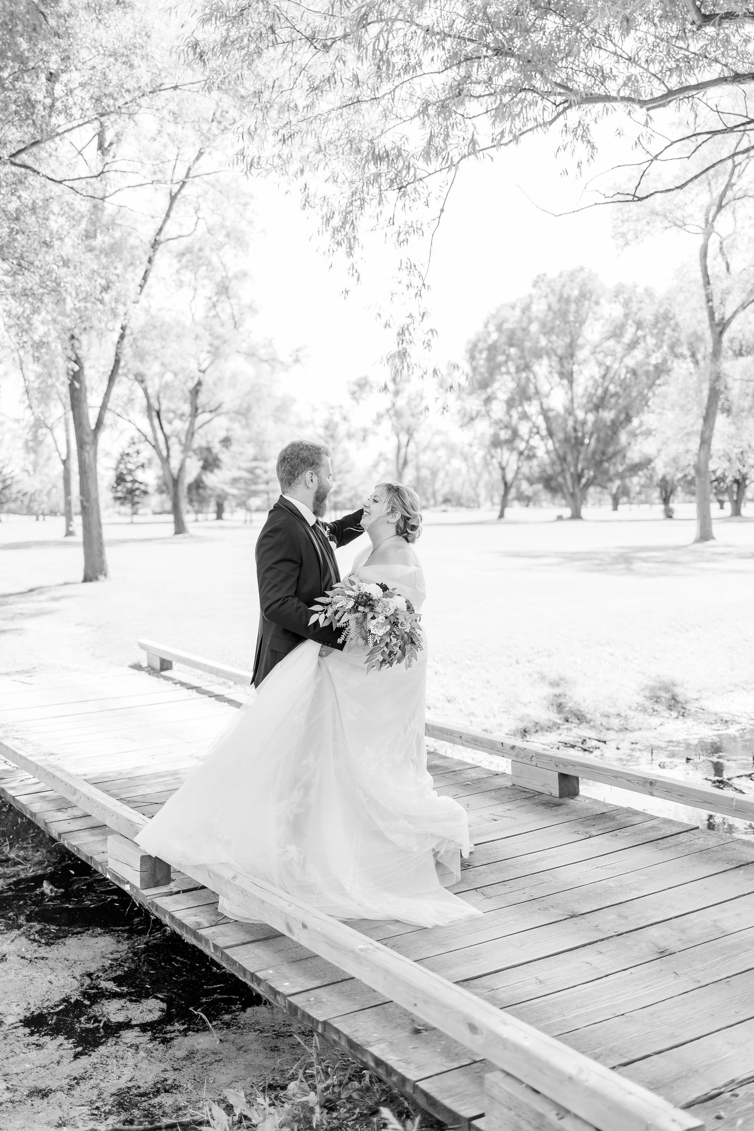 Black and white wedding photo on rustic wooden boardwalk surrounded by trees.