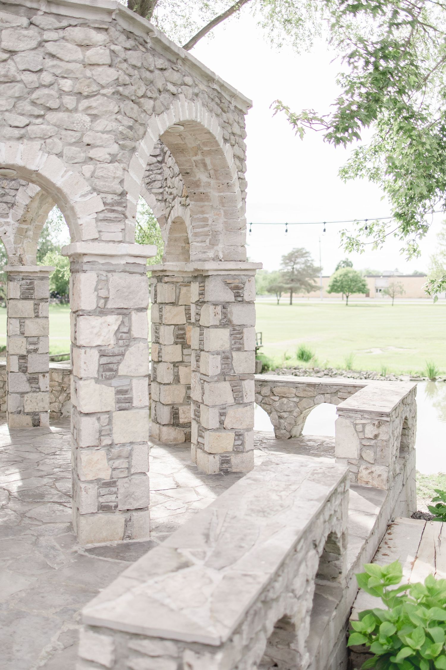 Rustic stone archway with decorative columns and string lights in a garden setting.