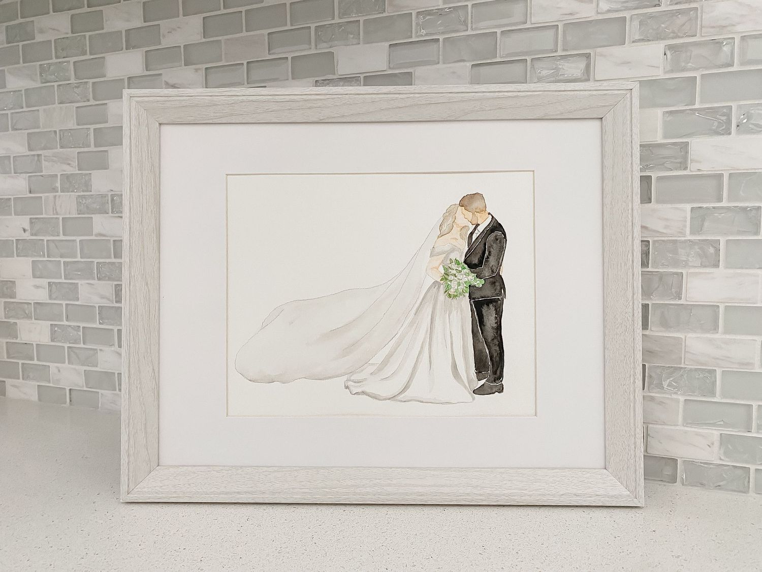 A delicate watercolor illustration of a bride and groom embracing, displayed in a white frame against a gray brick wall.