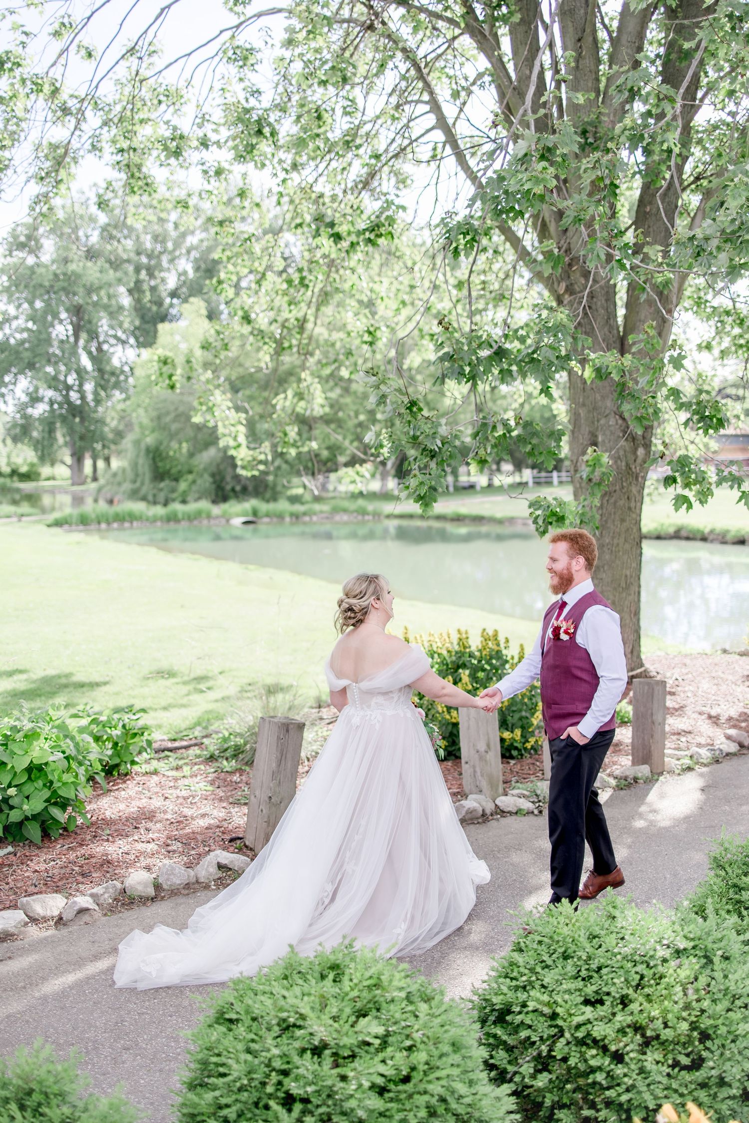 Newlyweds walk along a garden path surrounded by lush greenery and trees.