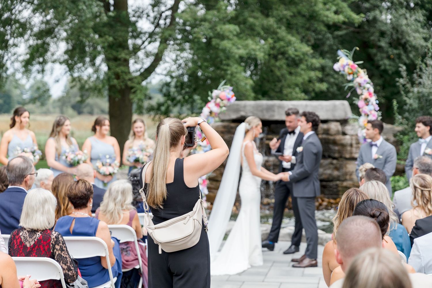 A photographer captures an outdoor wedding ceremony from behind guest seating.