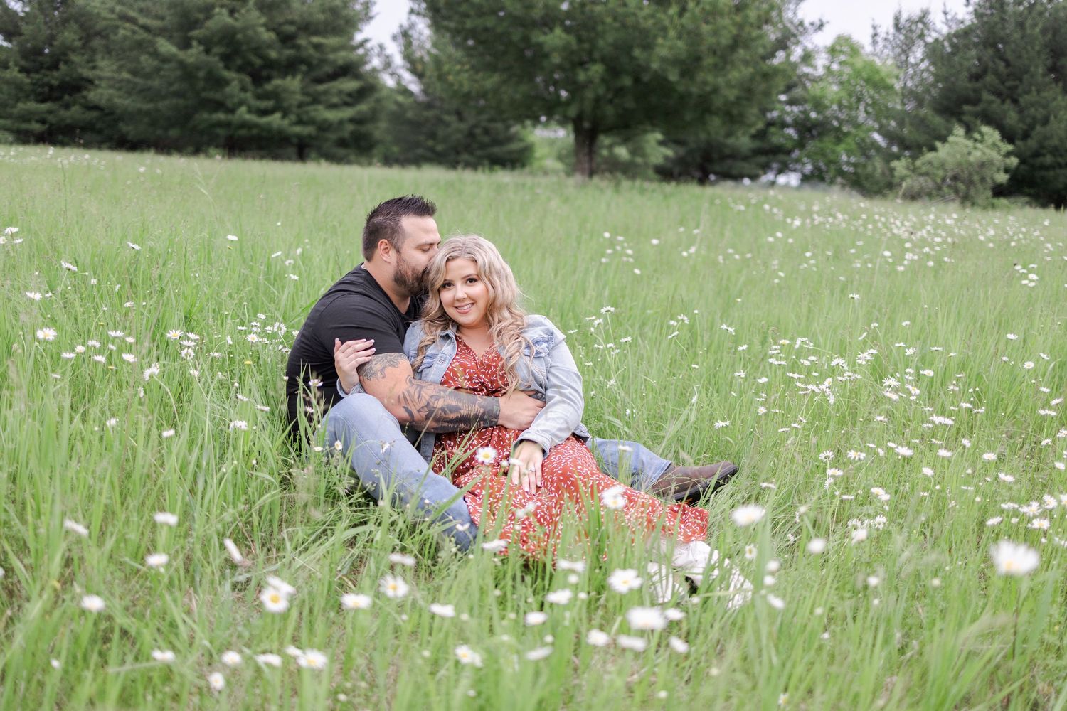 A couple shares an intimate moment while sitting in a serene meadow with pine trees.