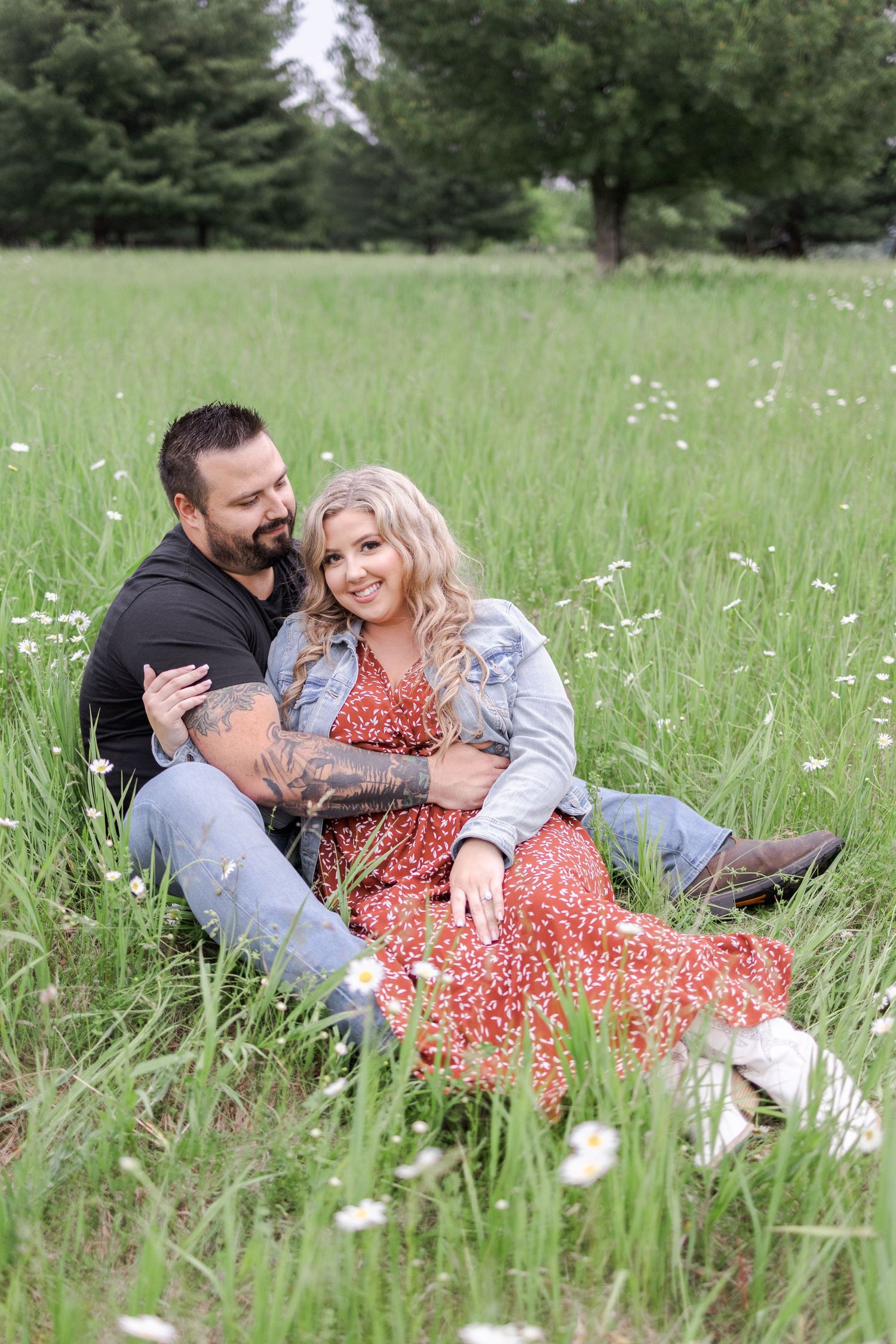 A couple sits together in a wildflower meadow with evergreen trees in the background.