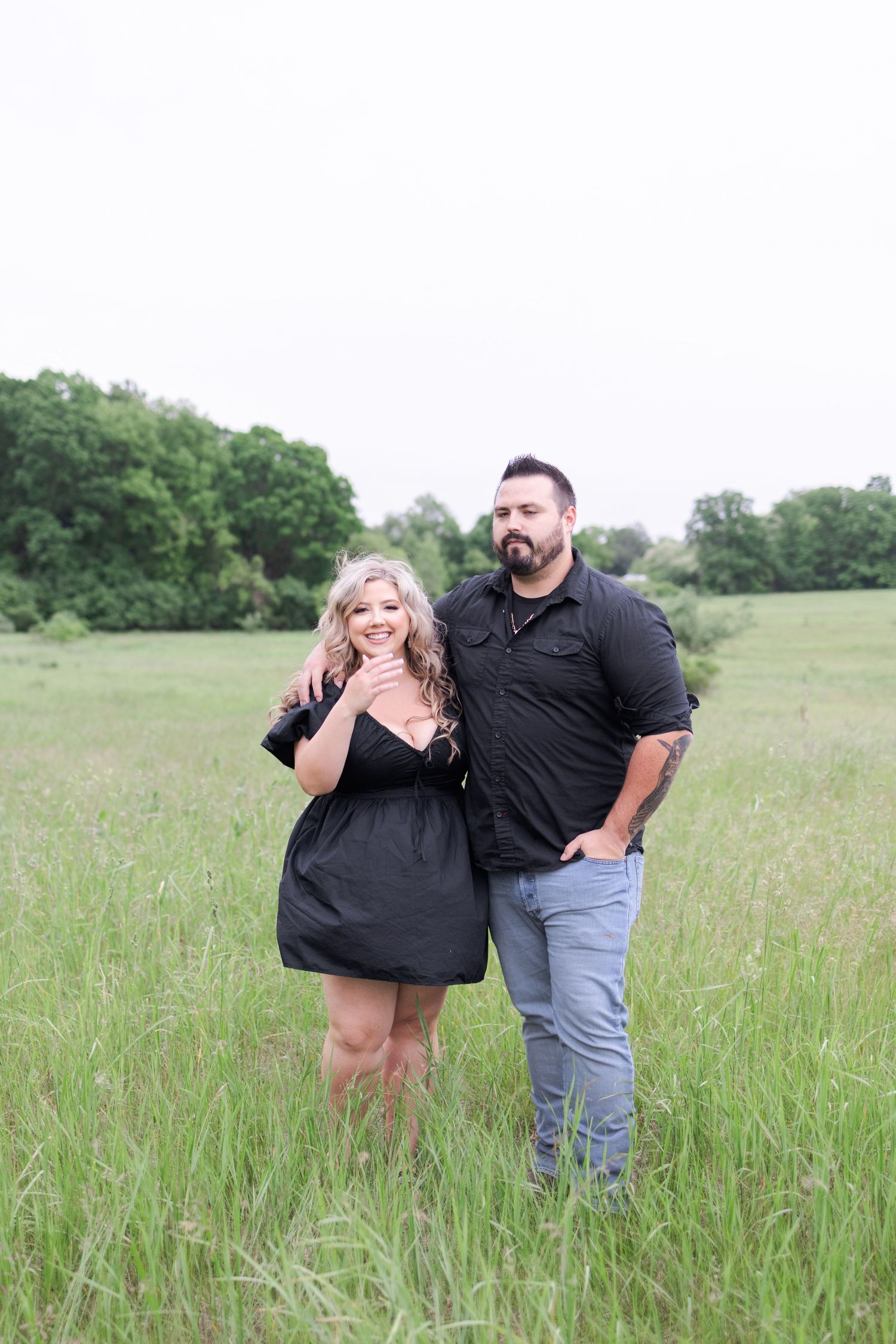 A romantic couple in black outfits poses playfully in a green meadow field.