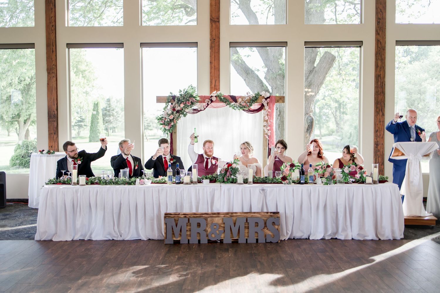 Long wedding reception head table setup with white linens and floral decorations in bright venue space.