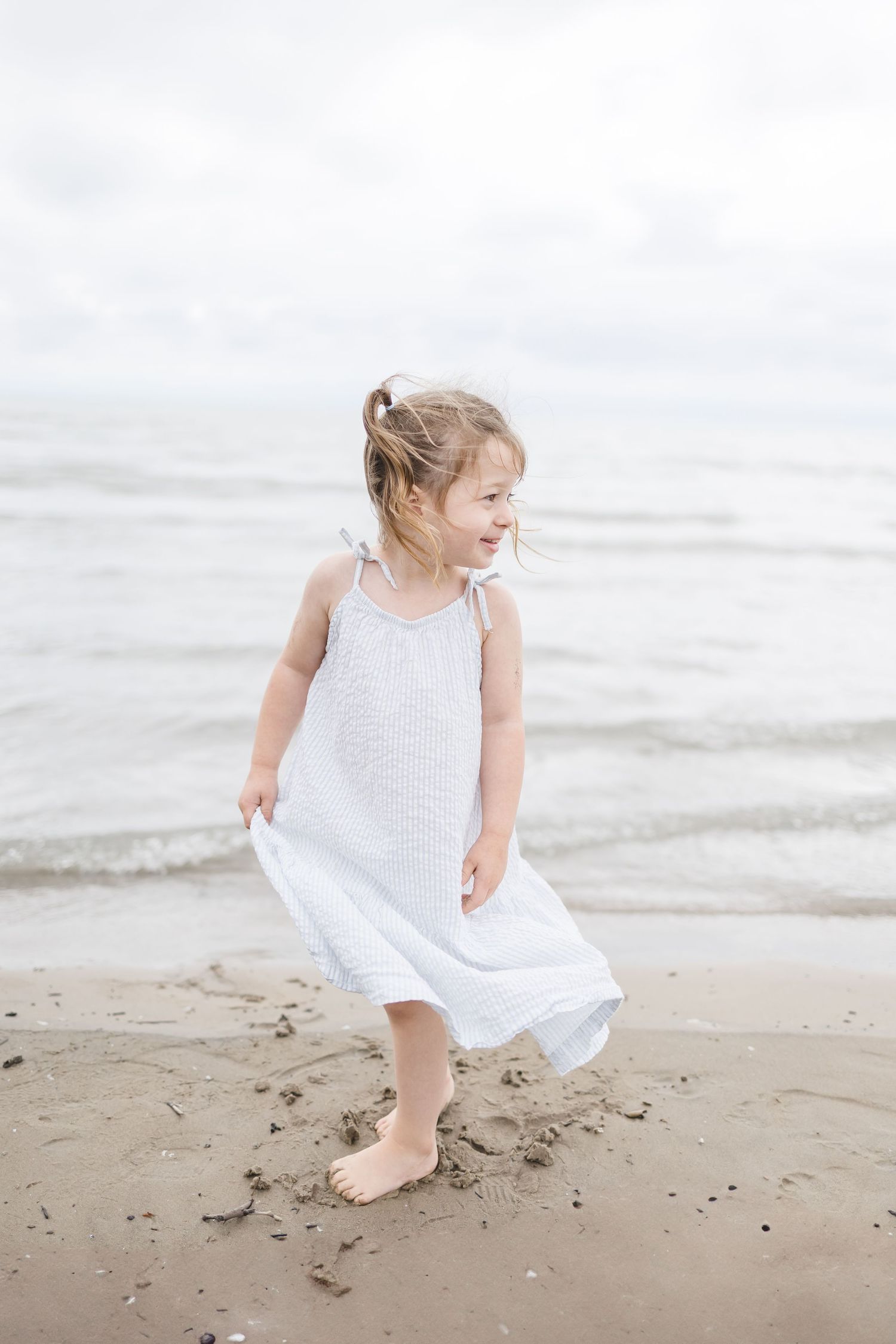 Young child in white dress playing and running on sandy beach.