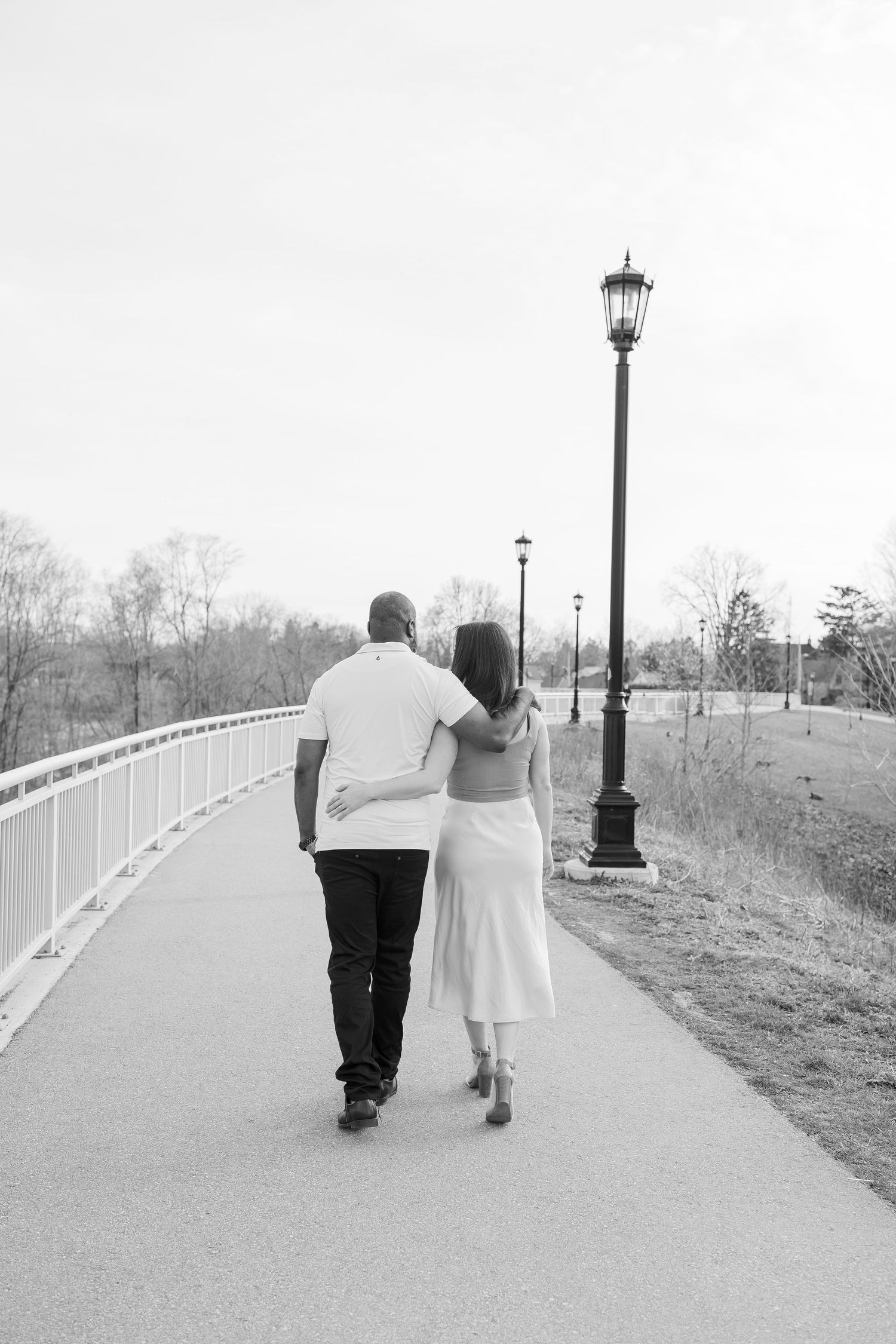 A couple takes a romantic stroll along a path with vintage lampposts in black and white.