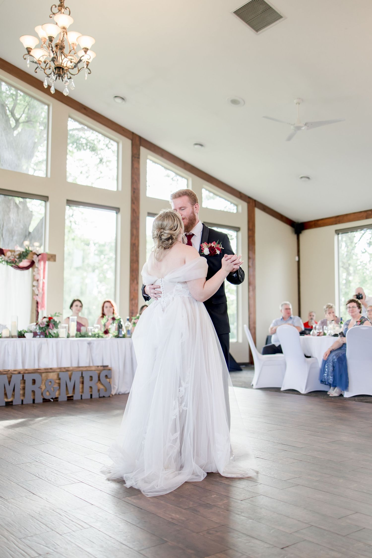 Wedding couple sharing first dance in spacious reception venue with wood floors and large windows.