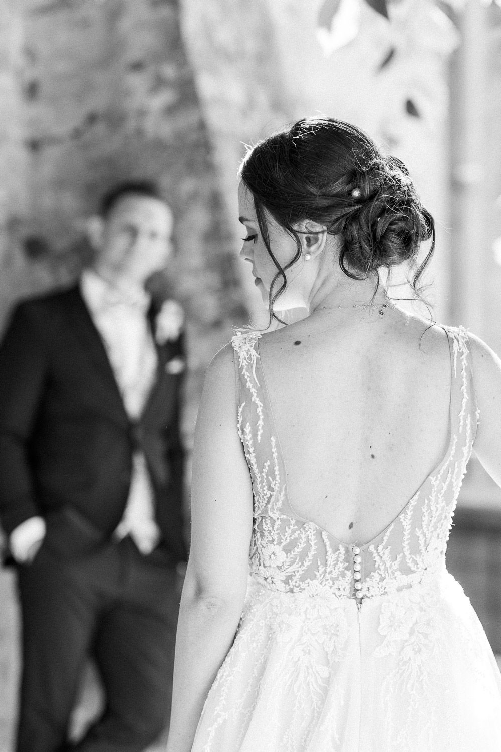 Black and white photo showcasing detailed back of wedding gown with groom in background.