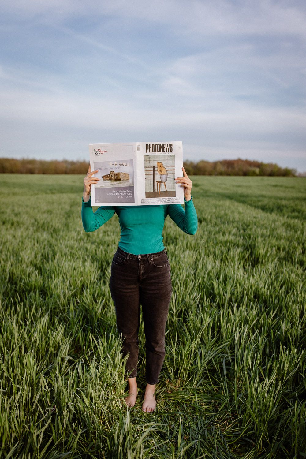 A person in a green top and jeans reads a magazine while standing barefoot in a grassy field.