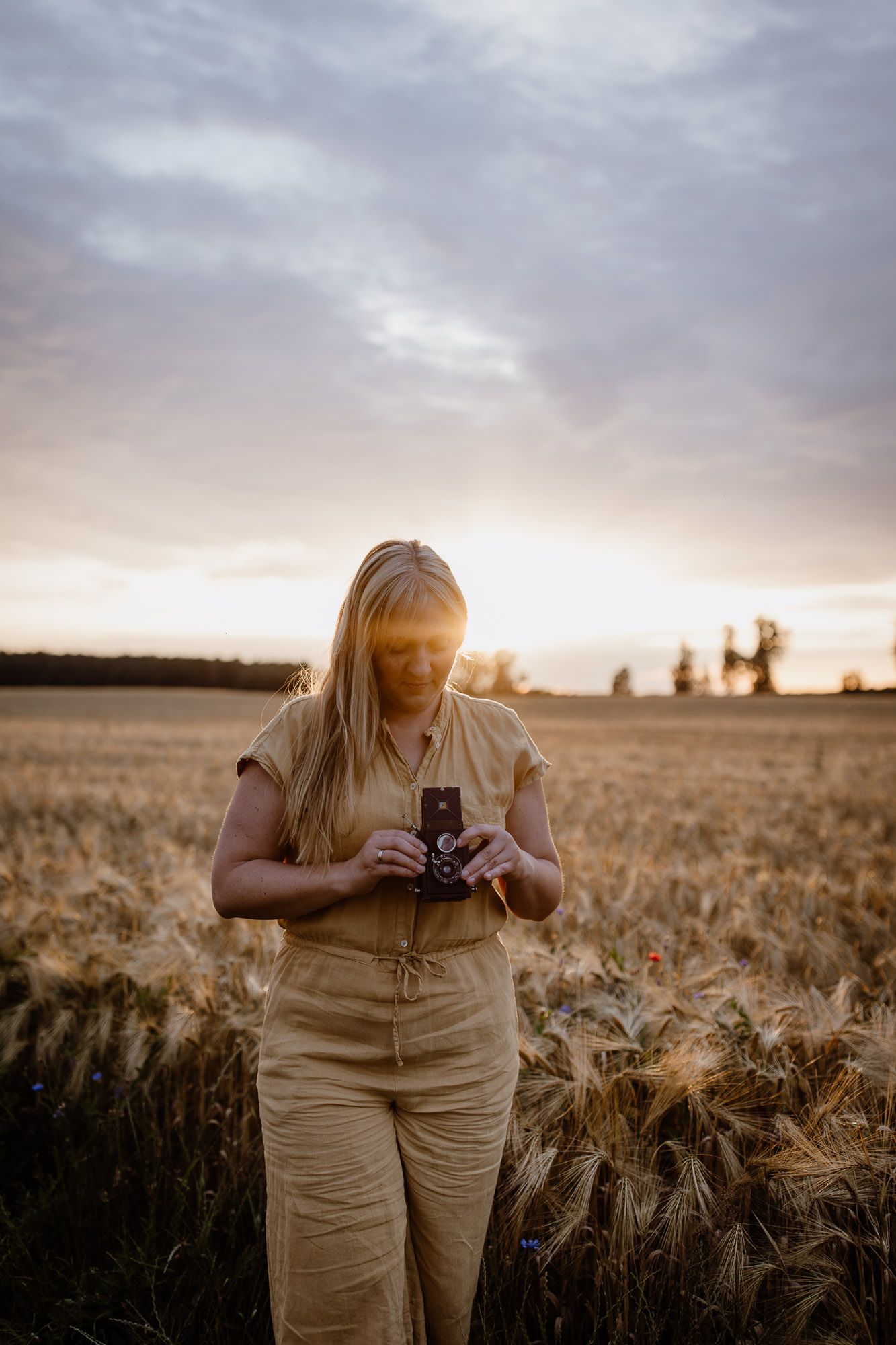 Person in casual attire photographs with vintage camera in wheat field during golden hour.