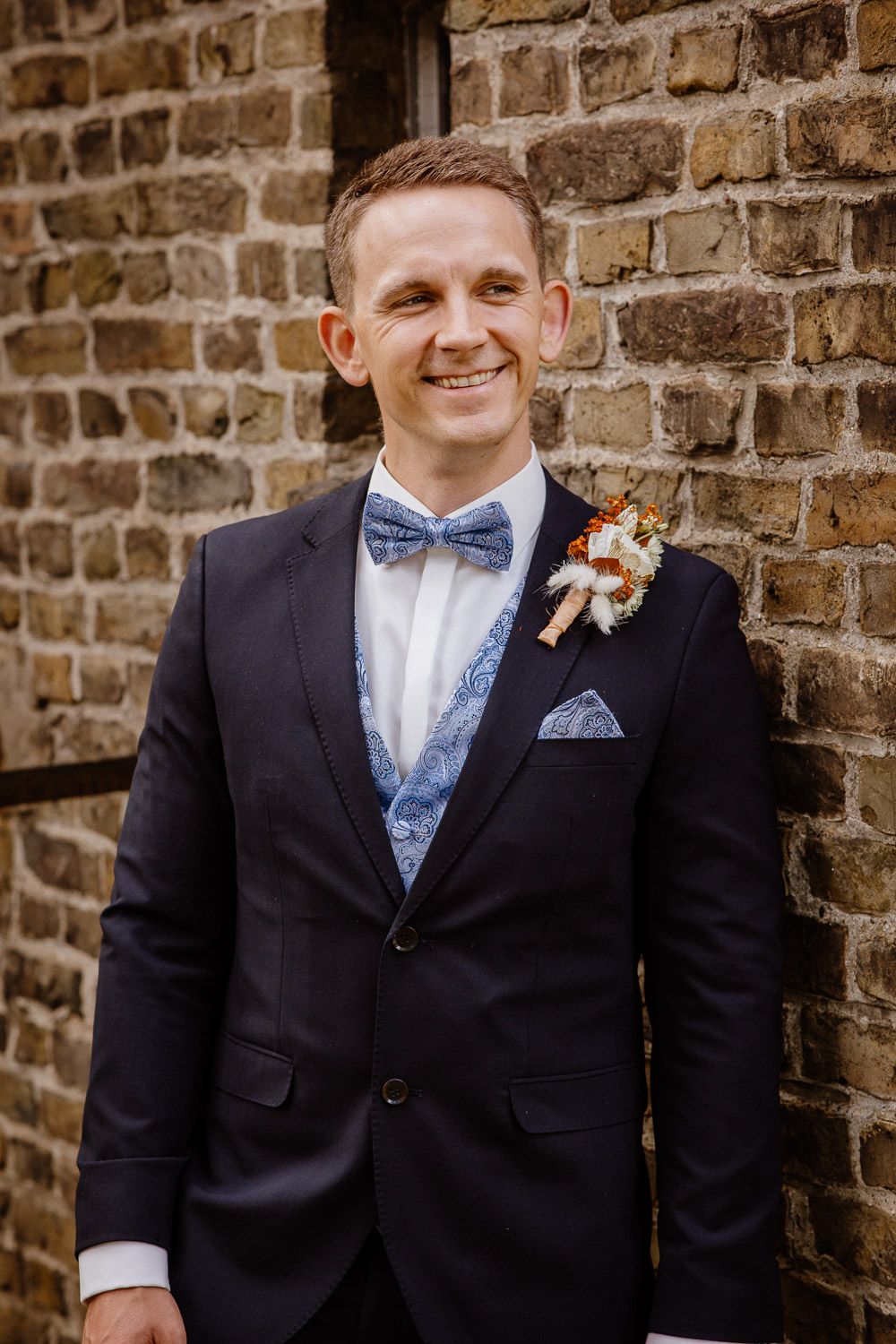 Groom in navy suit with bow tie and boutonniere smiling against brick wall background.