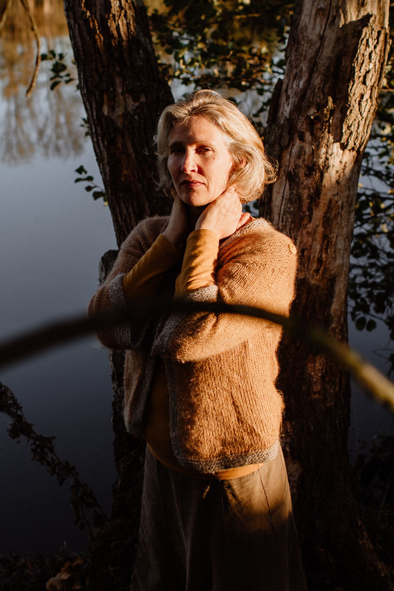 A person in a brown sweater stands between tree trunks at sunset, with water visible in the background.