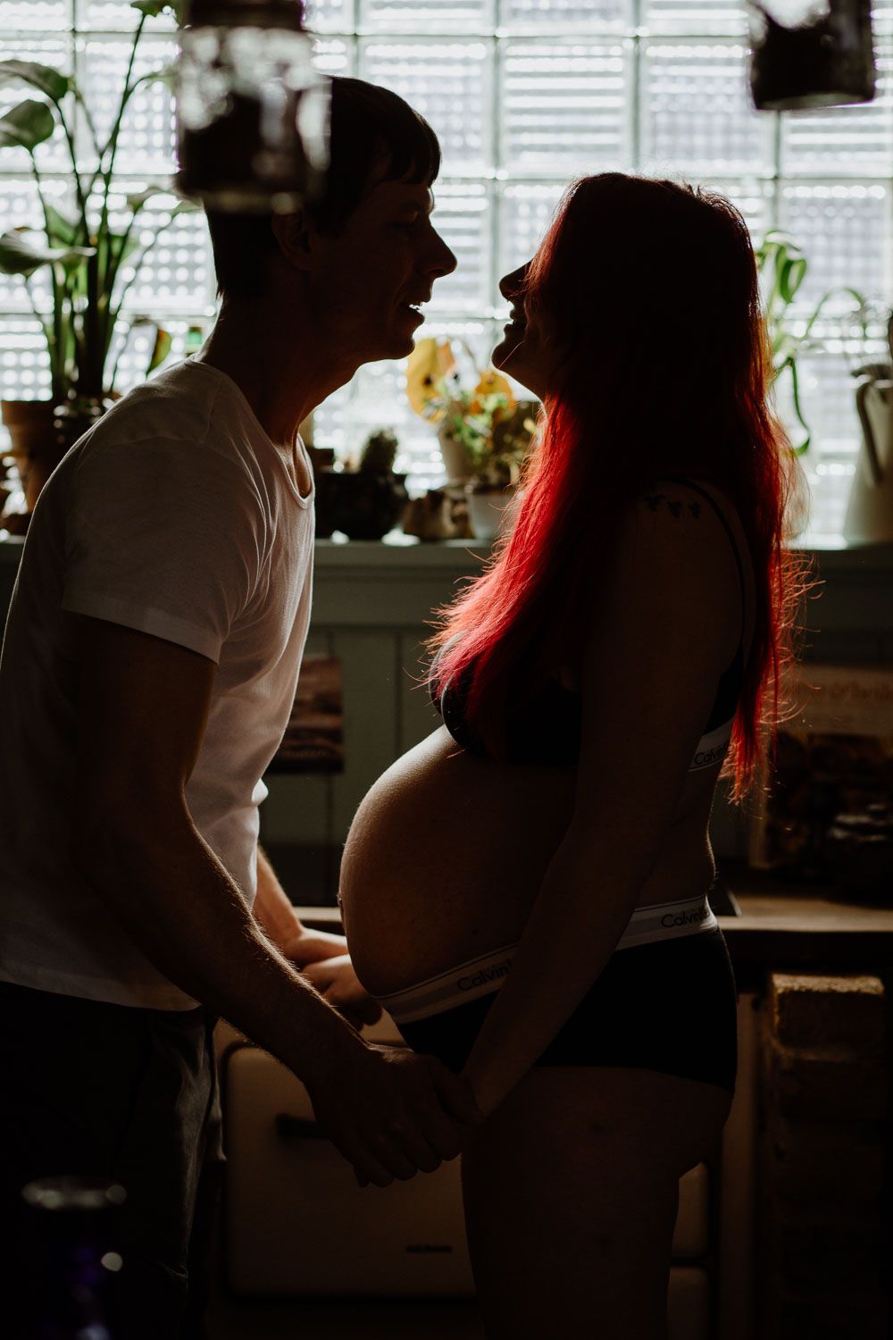 Silhouette of an expectant couple standing by a window with houseplants.