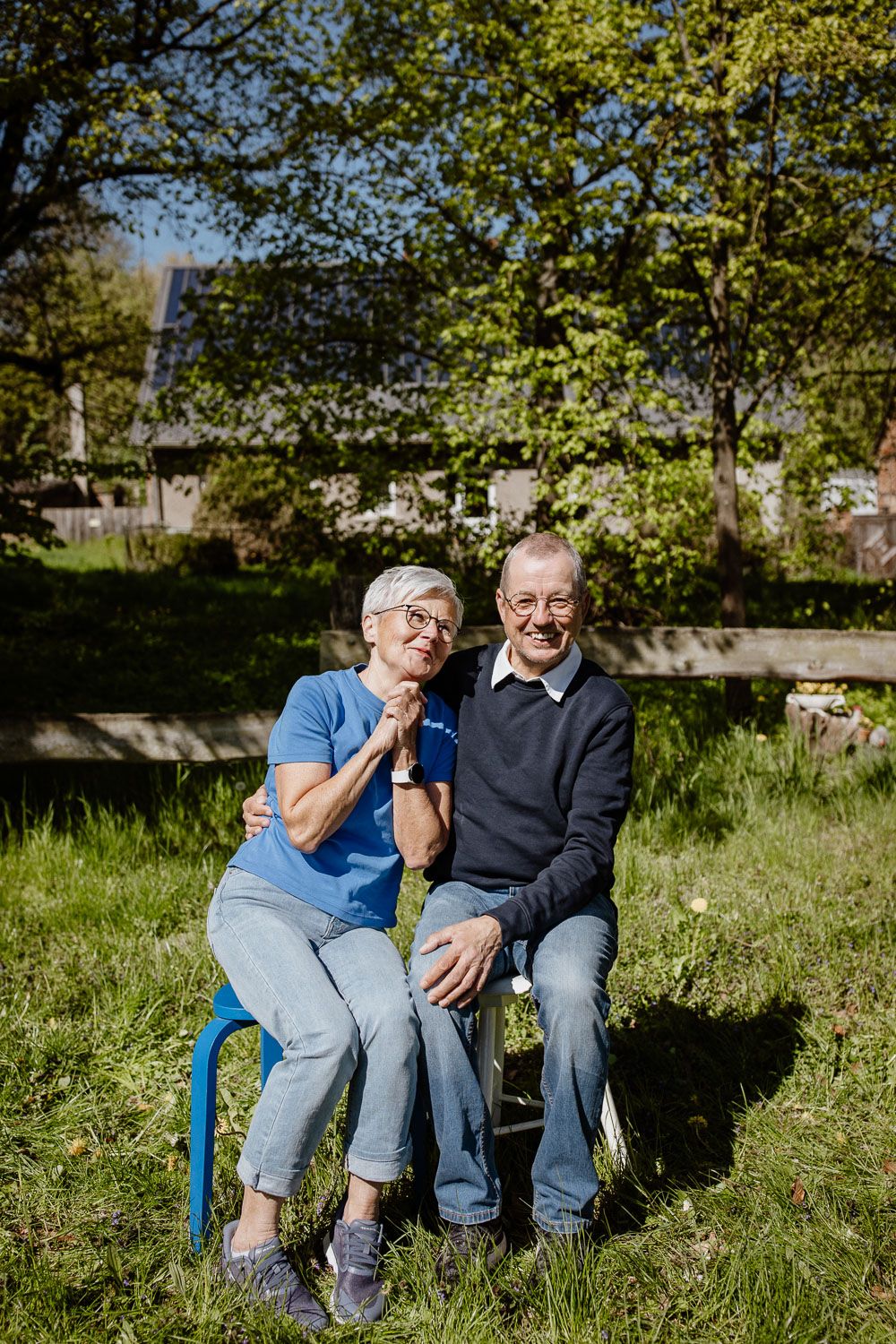 An elderly couple sits together on a wooden bench in a sunny garden setting.