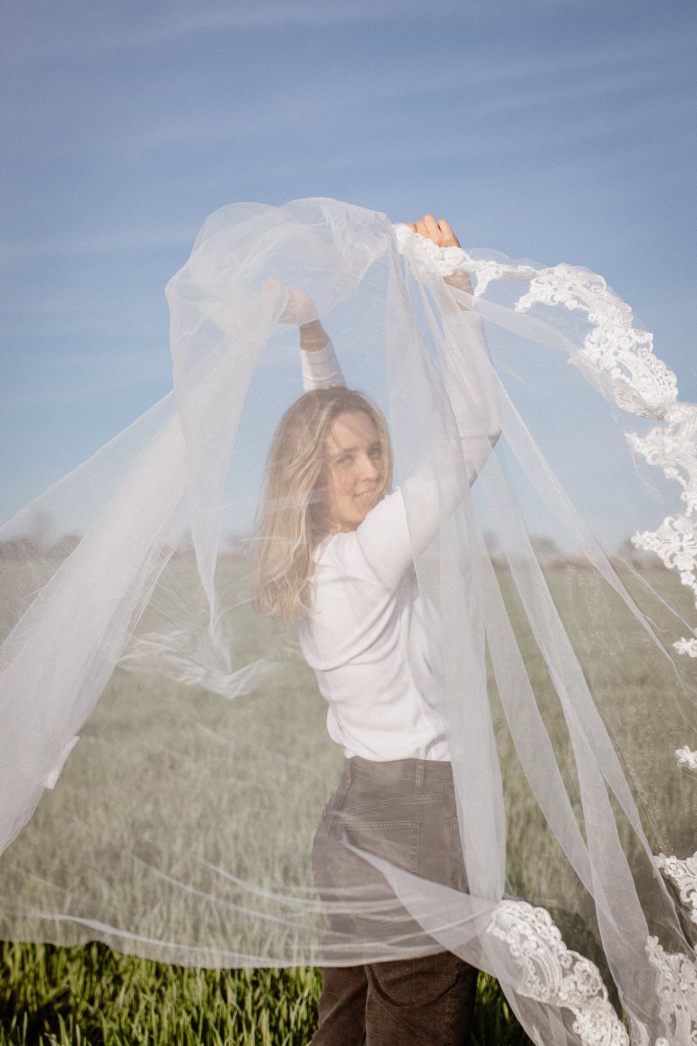 Person holding white lace veil overhead in sunny field with grass and blue sky.