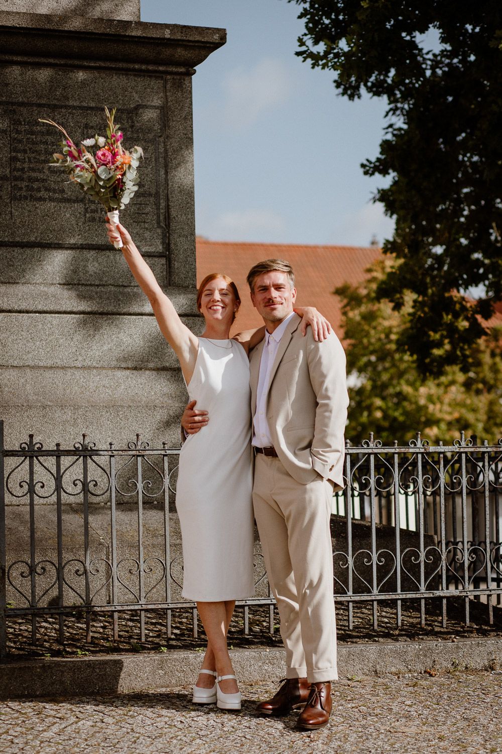 Wedding couple celebrating outdoors by ornate iron fence on sunny autumn day.