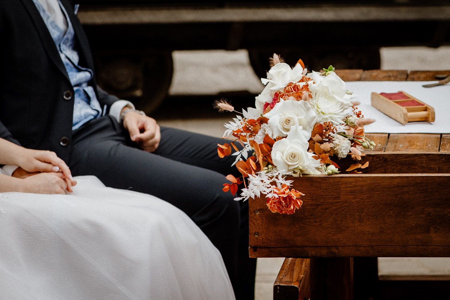 A wedding bouquet with white and coral flowers rests on a wooden bench next to formal attire.