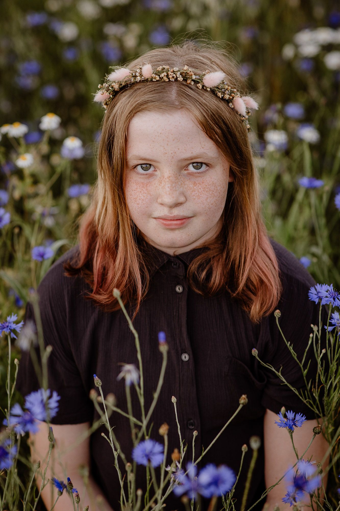 A delicate floral crown adorns red hair as the subject sits among vibrant blue cornflowers in a dark shirt.