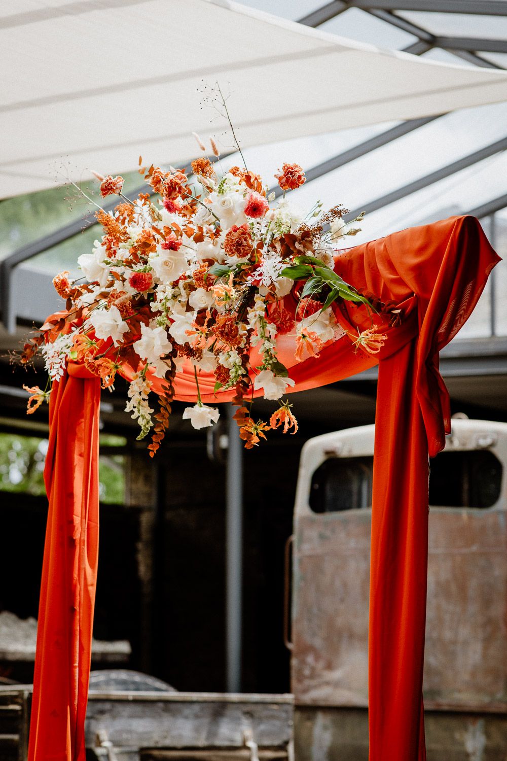 Wedding arch decorated with orange drapes and white floral arrangements against a brick wall.