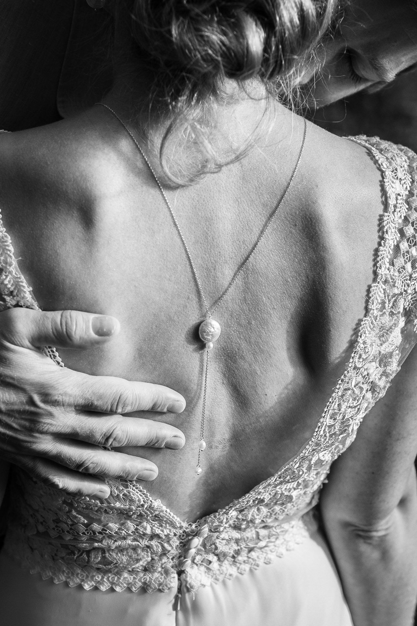 Black and white photo showing delicate lace back of wedding dress with a simple necklace pendant against bare skin.