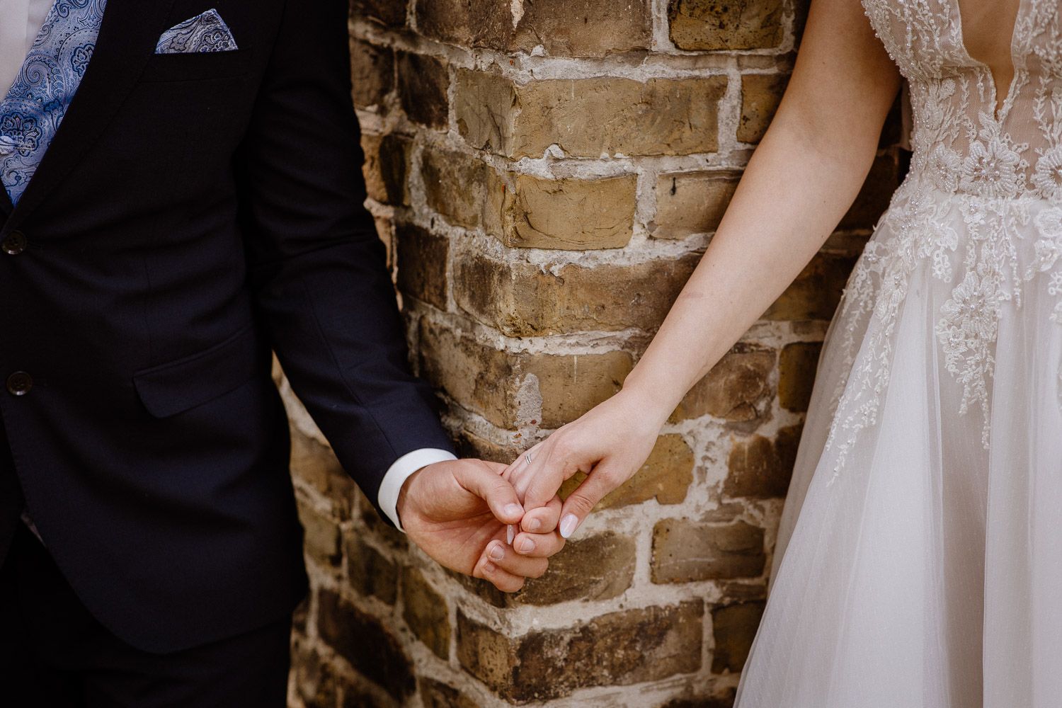 Close-up of couple holding hands showing wedding rings against rustic brick wall backdrop.