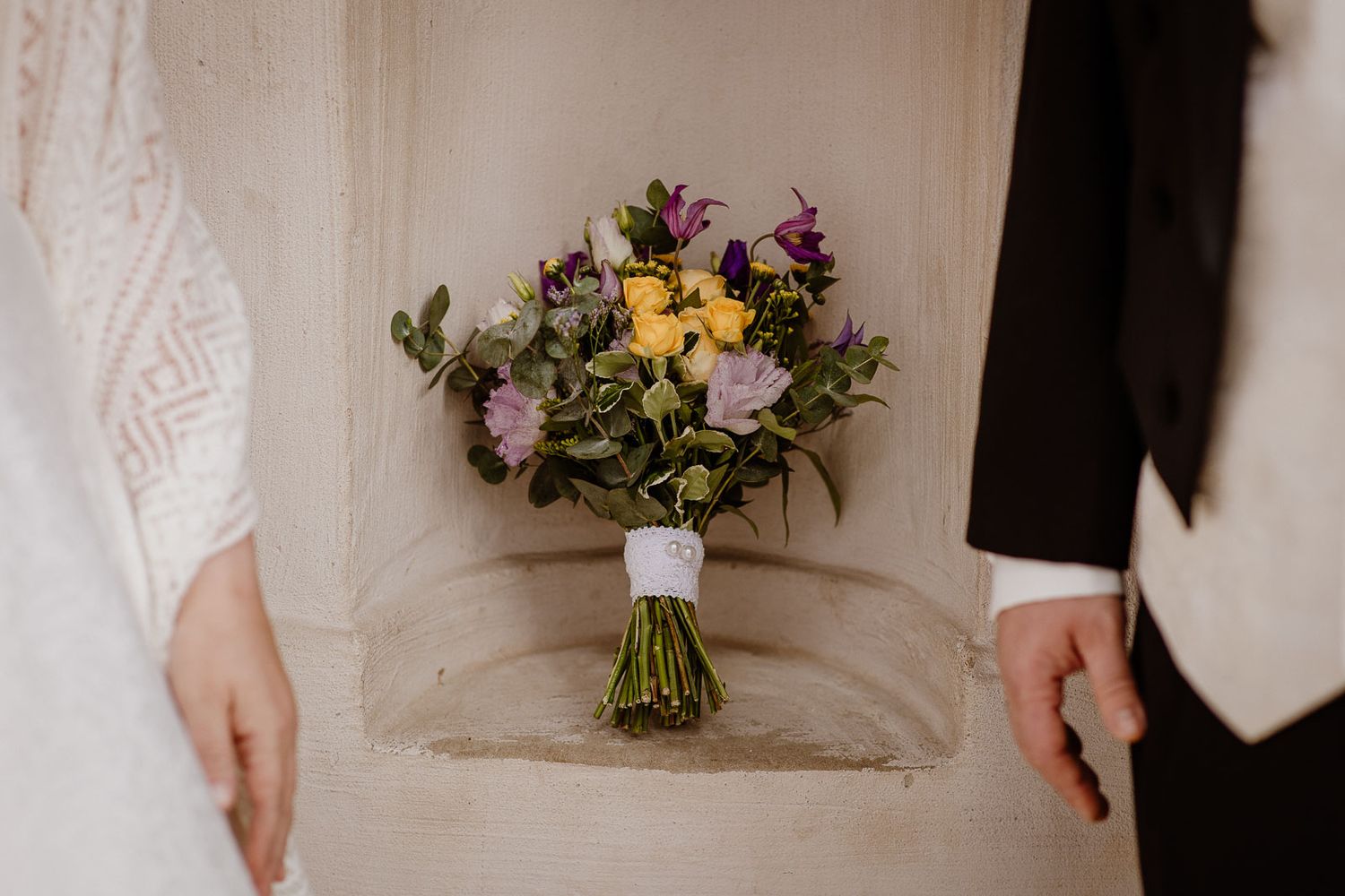 A wedding bouquet with yellow and purple flowers wrapped in twine being held between a white dress and black suit.