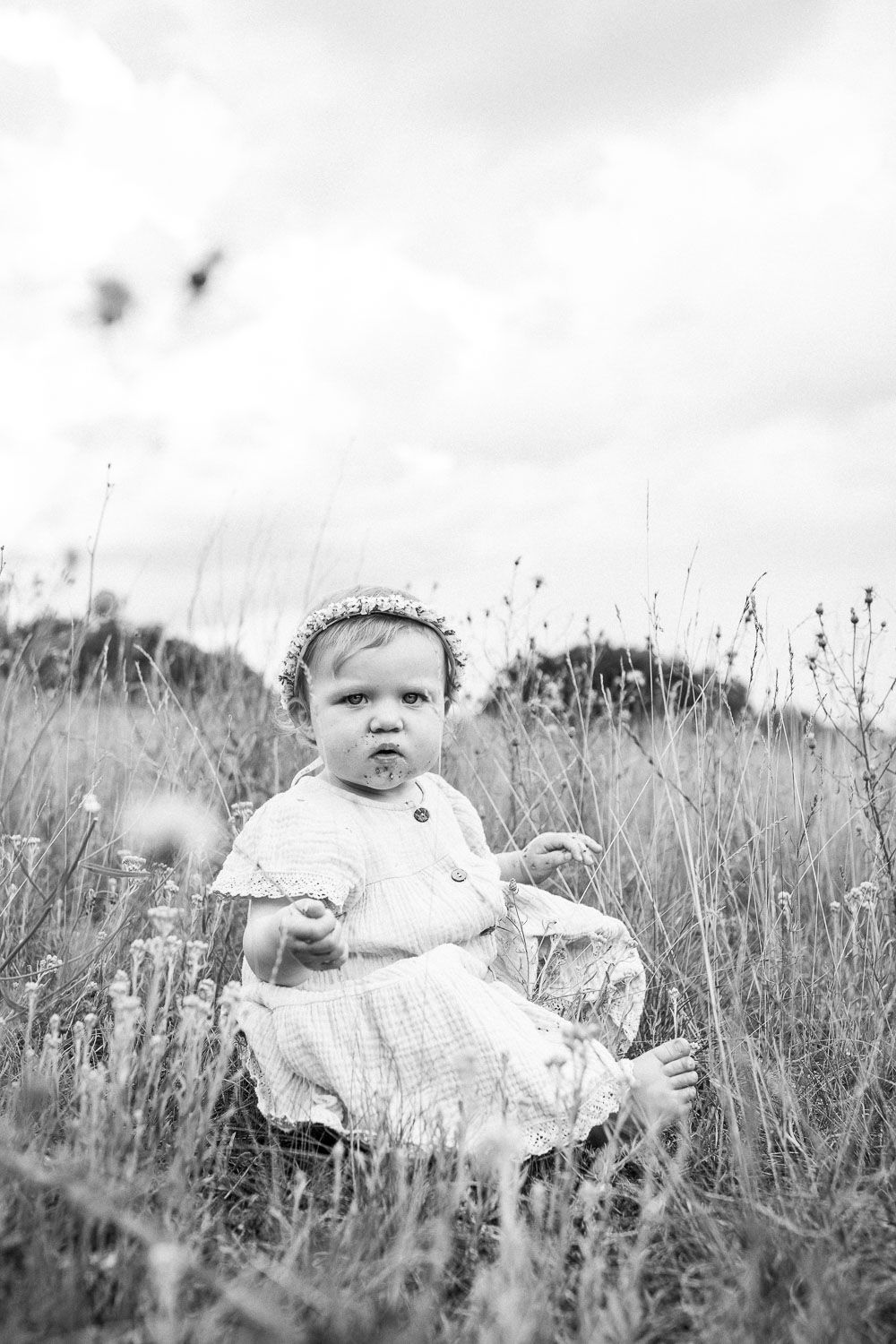 Black and white portrait of a child sitting in a wildflower field wearing a white dress.