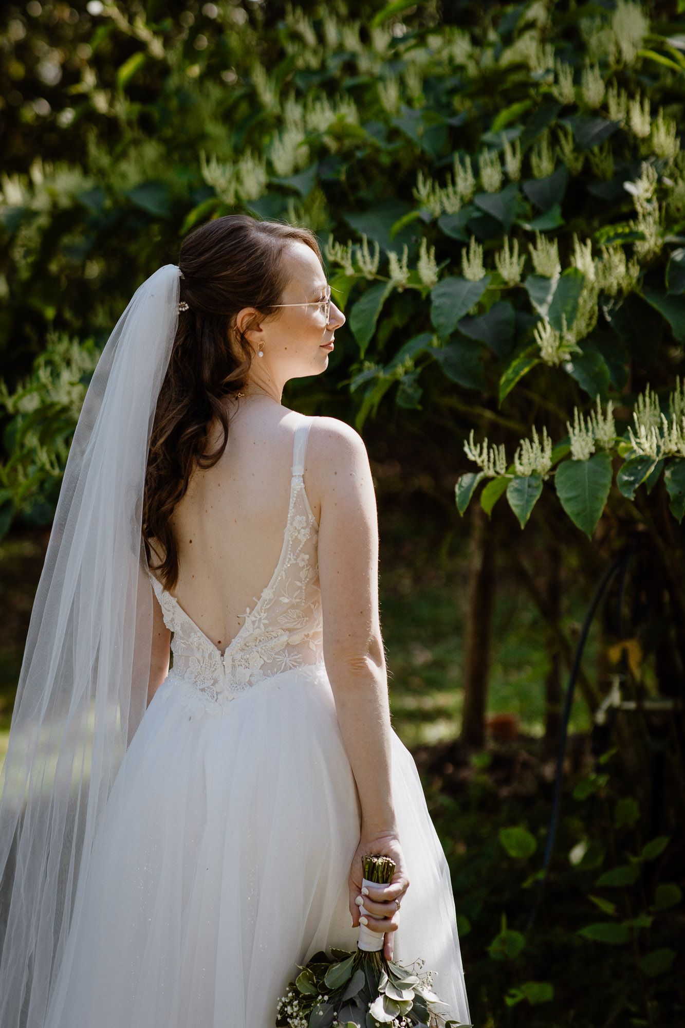 Back view of bride in white lace gown with flowing veil standing against lush greenery in garden setting.