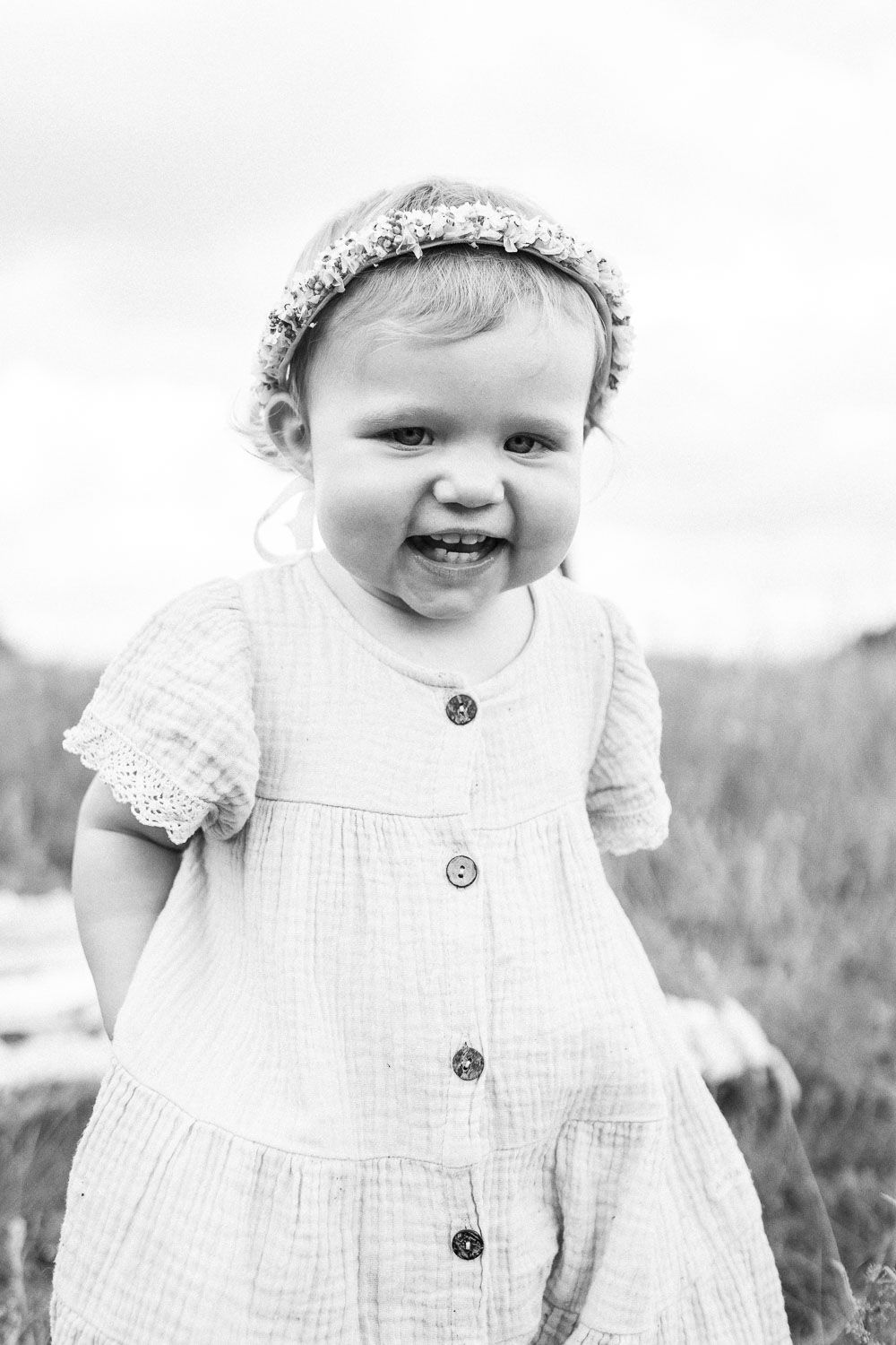 A baby in a white dress and floral crown smiles joyfully in black and white.