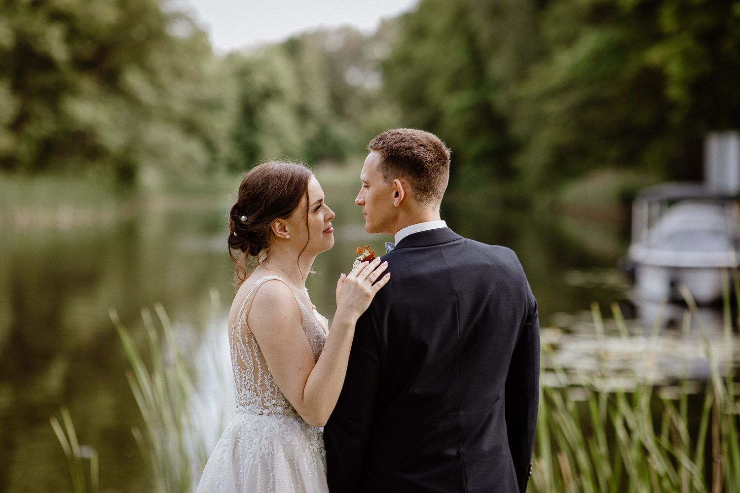 A romantic wedding couple embraces by a serene lake with lush greenery in the background.
