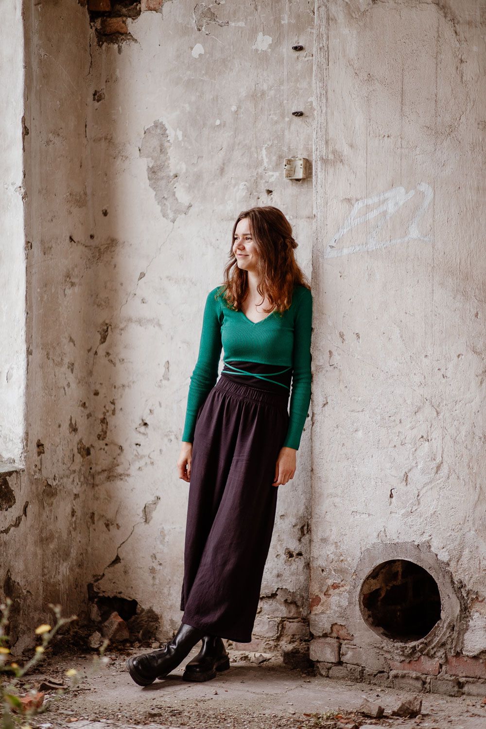 A woman in a green sweater and long brown skirt leans against an industrial concrete wall in an artistic pose.