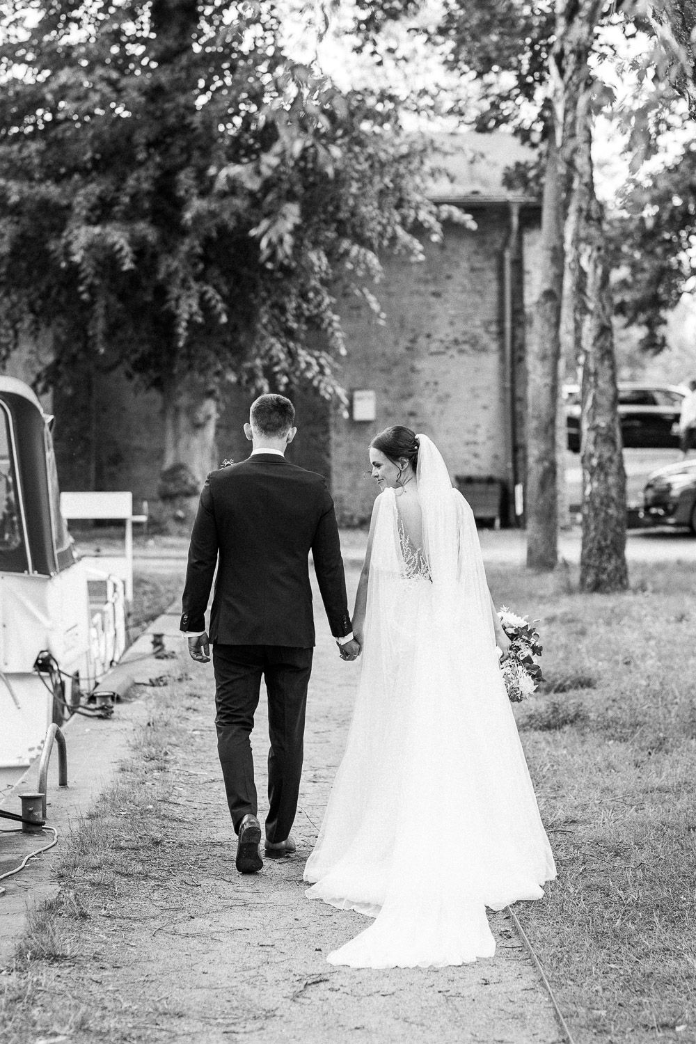 Black and white photo of newlyweds walking hand in hand down a tree-lined path.