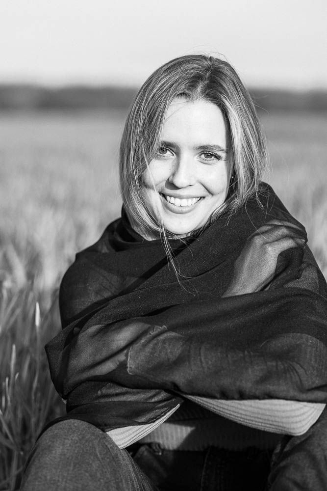 Black and white portrait of someone wrapped in dark scarf smiling while sitting in outdoor field setting.