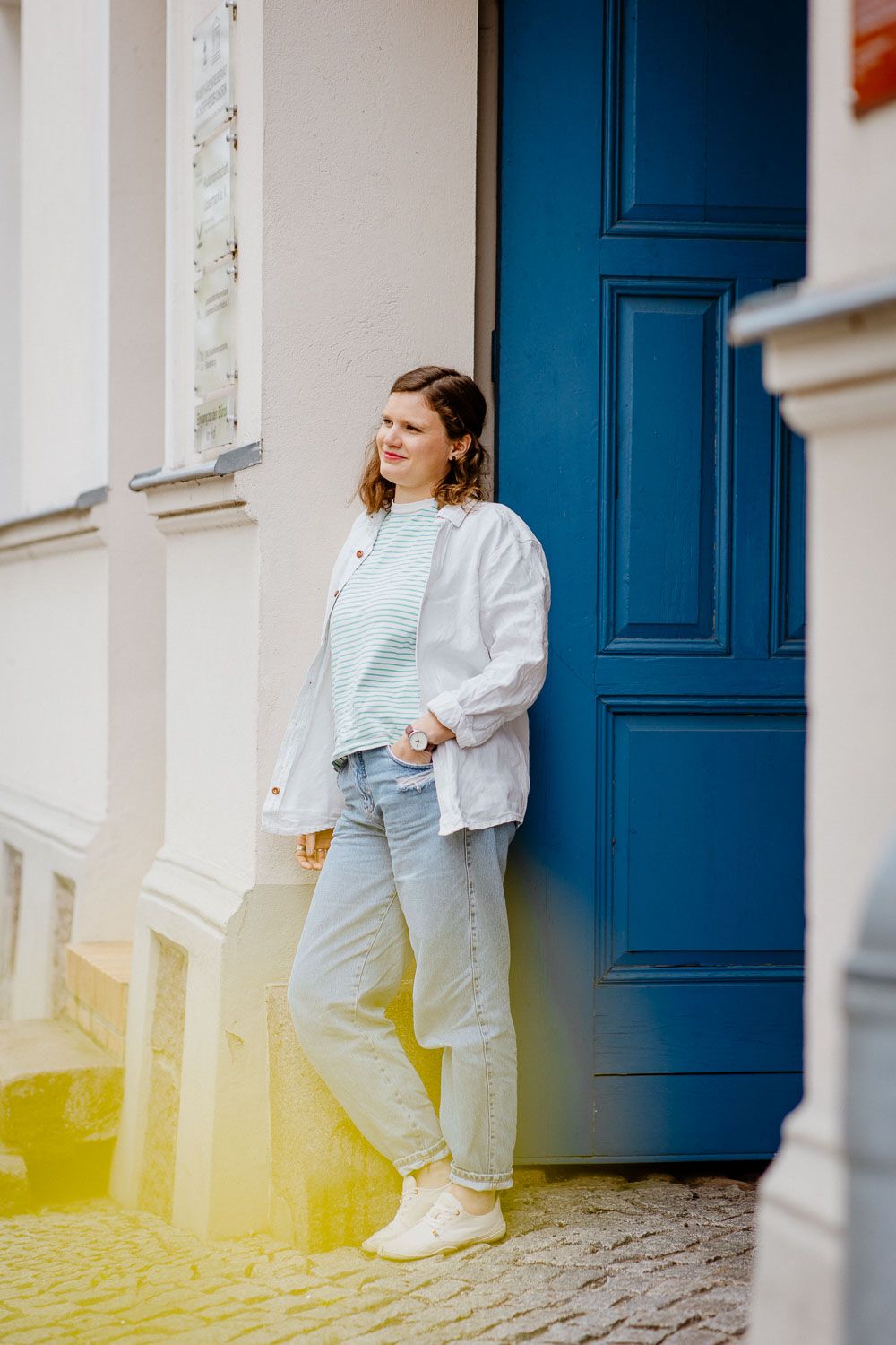 Woman in casual white outfit leaning against blue door in natural sunlight.