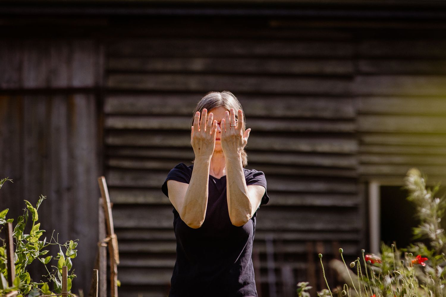 A person covers their face with both hands while standing in a garden.