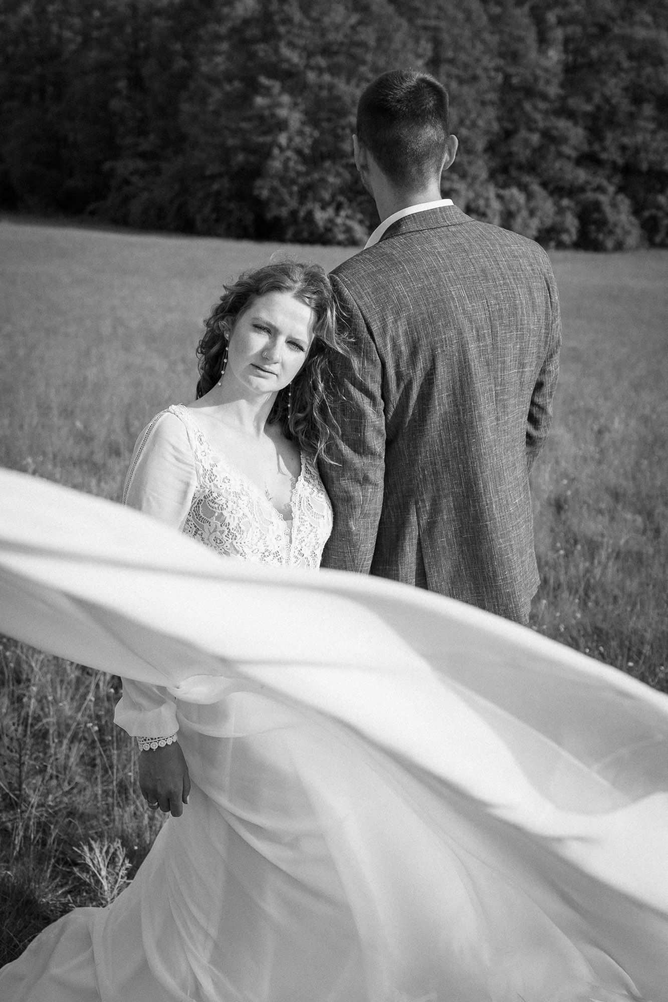 Black and white wedding photo showing a bride in flowing white dress and groom in suit standing in a grassy meadow.