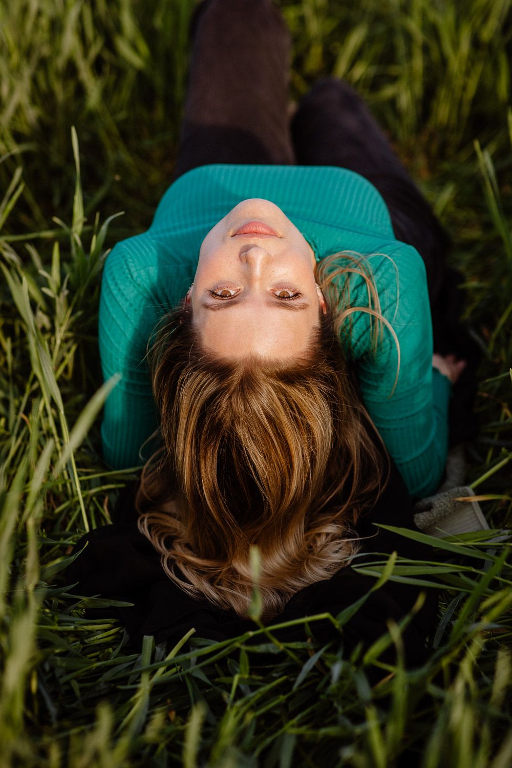 Woman in teal sweater lying in tall grass looking upward at camera.
